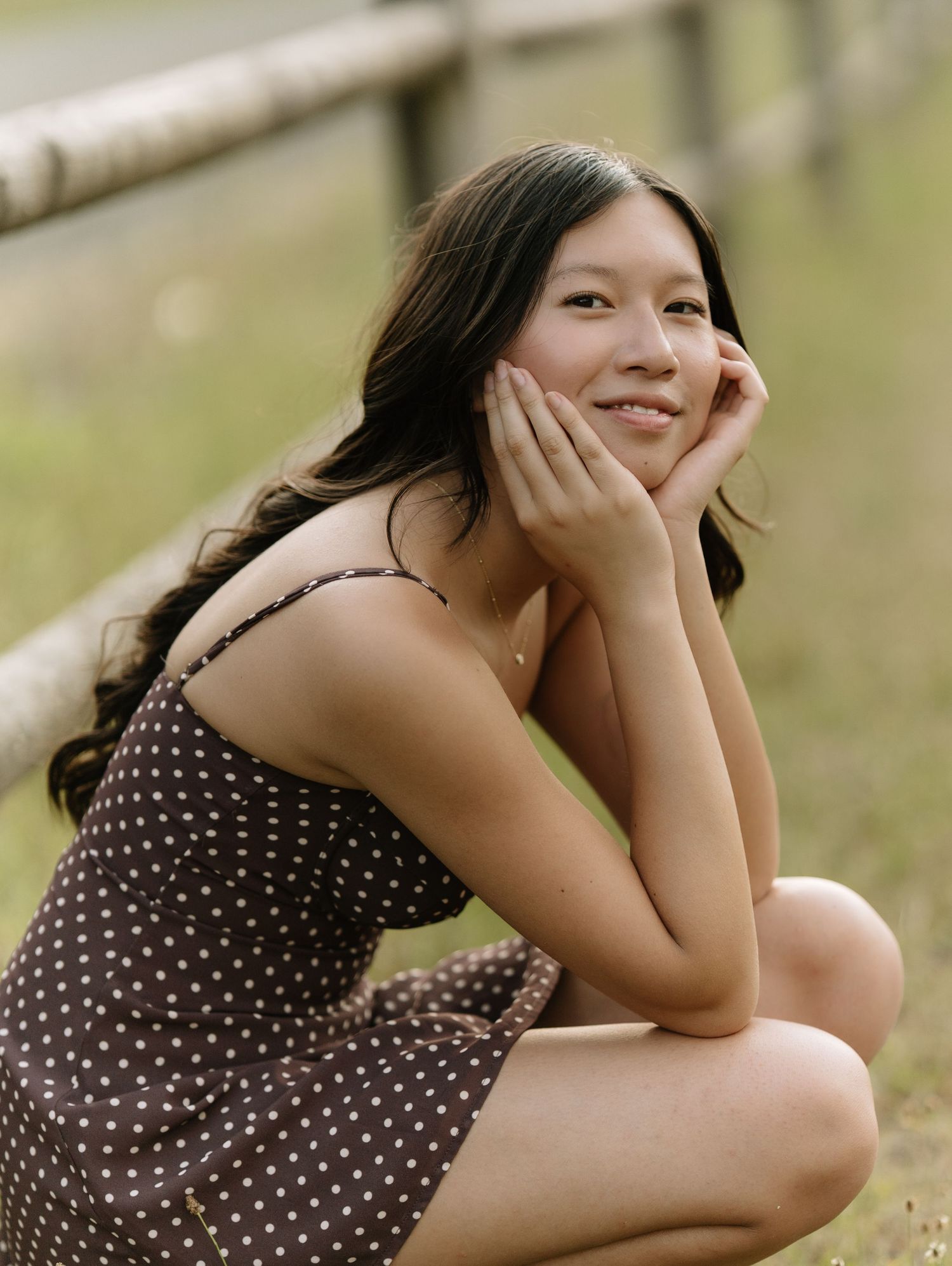 Smiling woman in a brown polka dot dress rests her chin on her hands while crouching beside a rustic wooden fence.