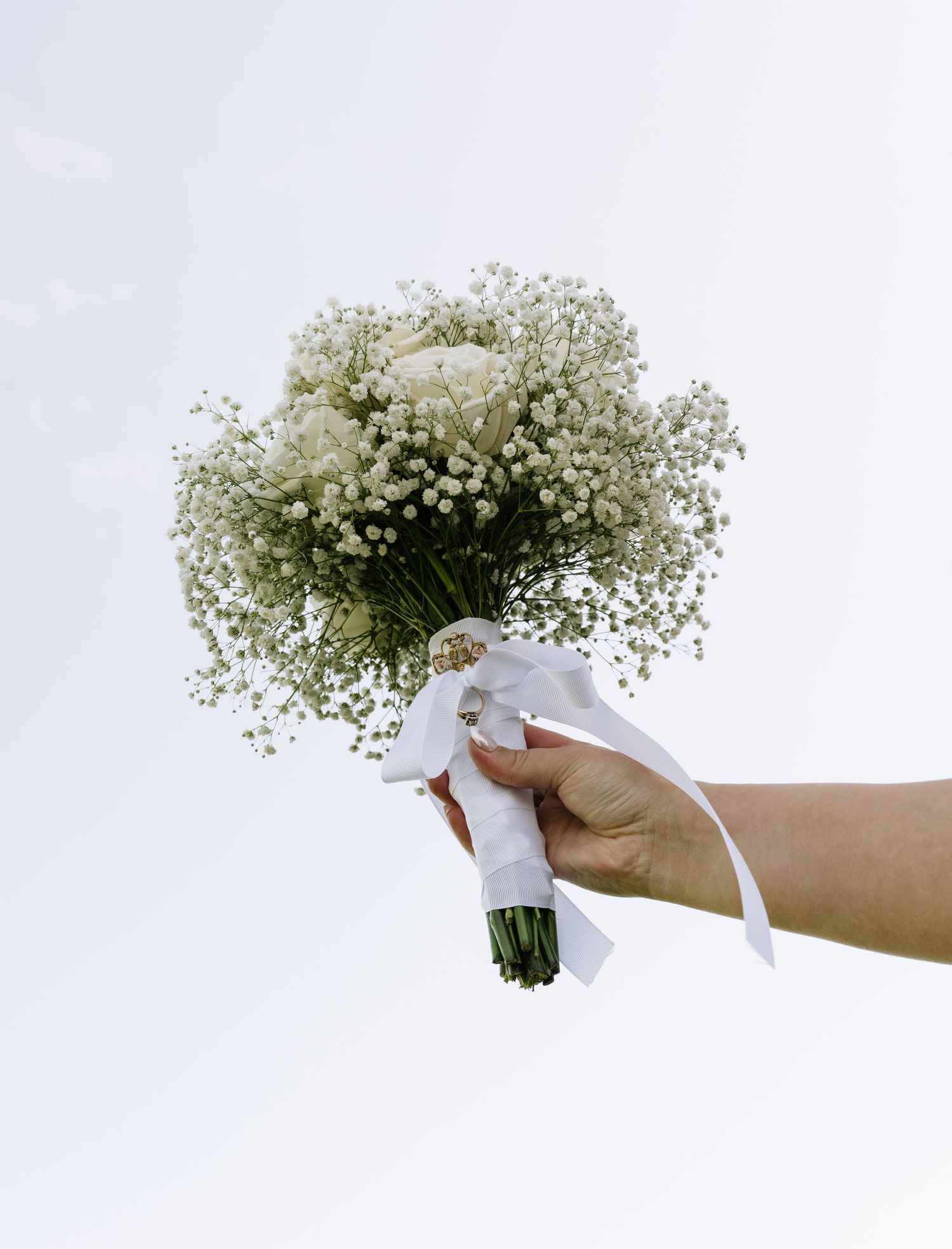 Hand holding a wedding bouquet of white roses and baby's breath tied with a white ribbon against a pale sky.