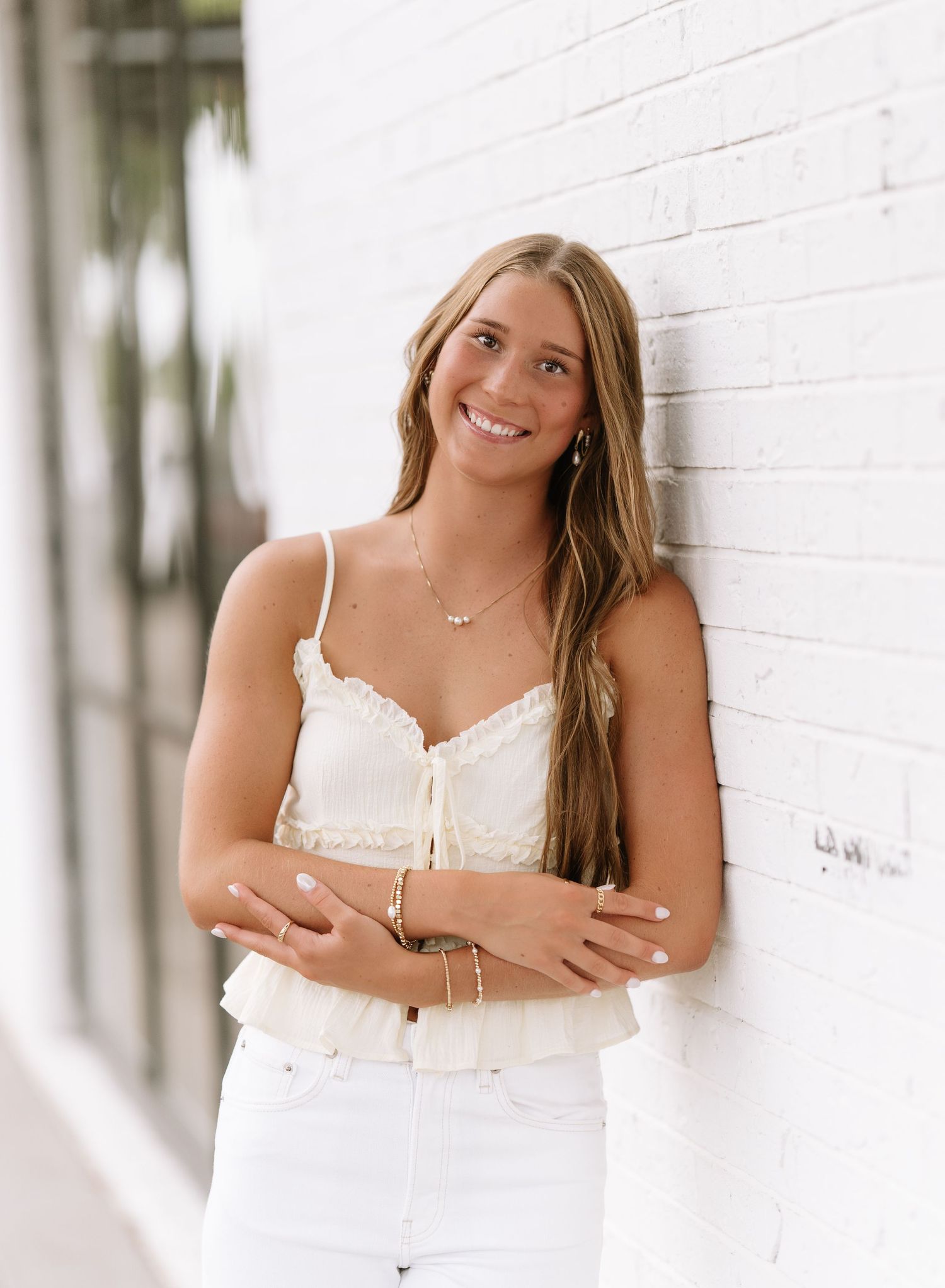 Smiling woman with long hair leans against a white brick wall in a cream top and white jeans for a senior portrait.