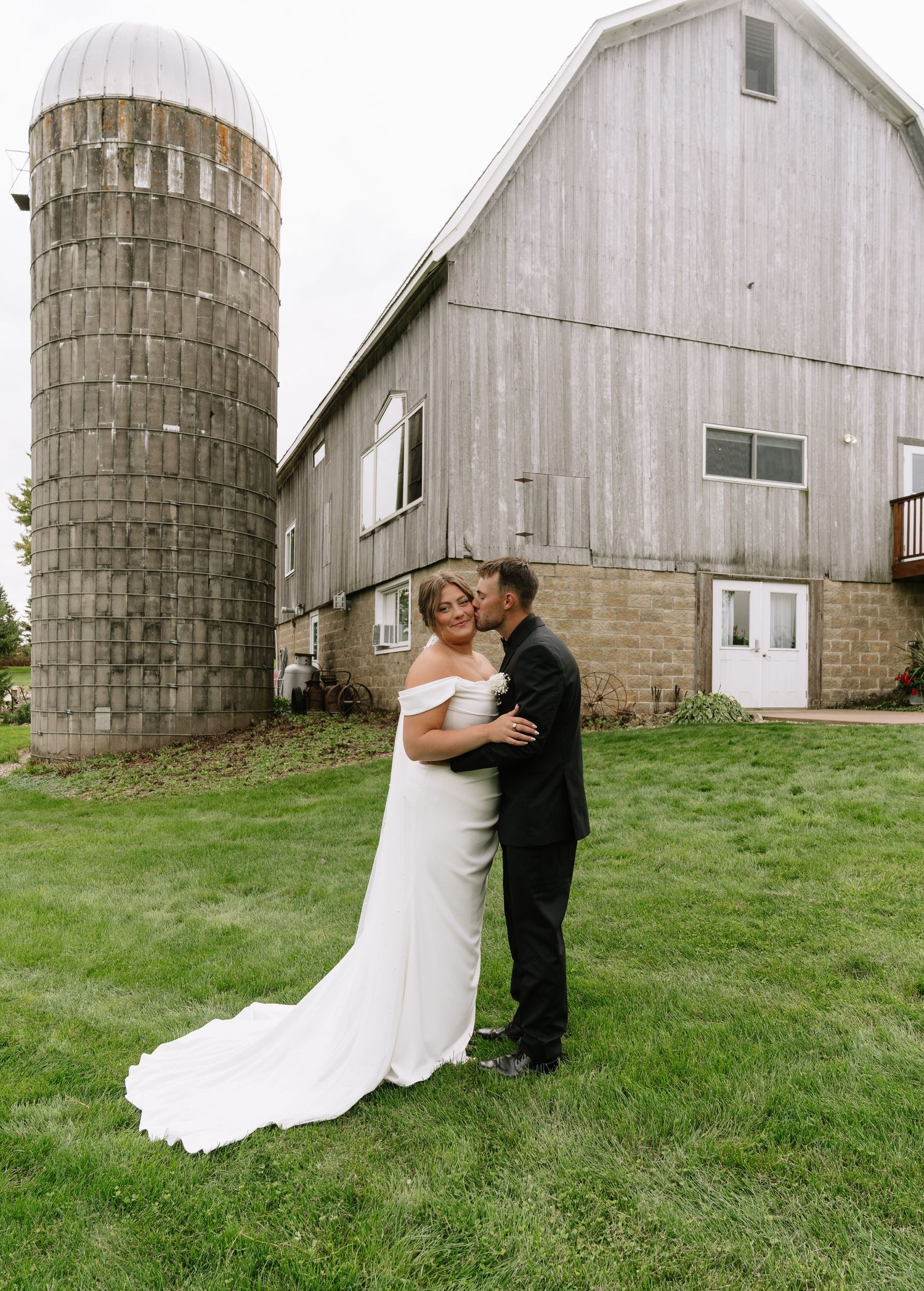 Bride in a white off-shoulder gown and groom in a black suit embrace on a lawn in front of a rustic barn and silo.
