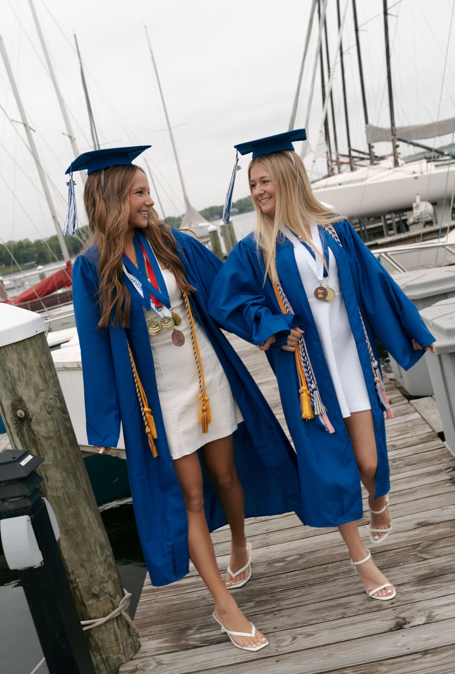 Two graduates in blue gowns, caps and honor cords walk arm in arm along a wooden marina dock with sailboats in the background.