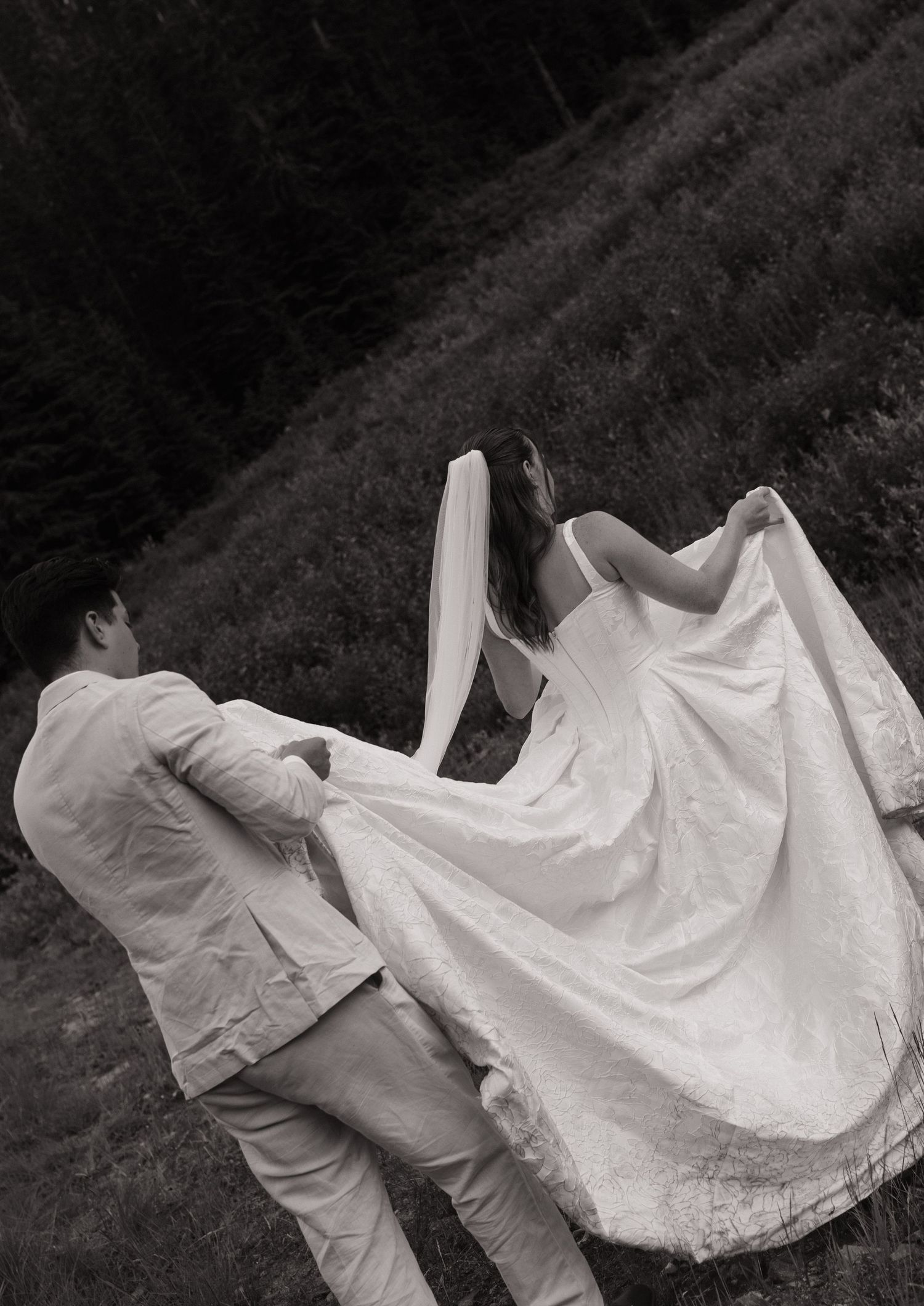 Black and white photo of a bride in a flowing satin gown holding her skirt while walking up a grassy hill with her groom.