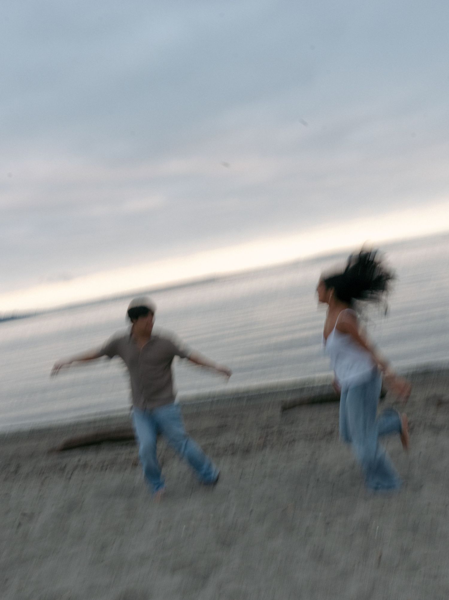 Couple in motion on a pebbly beach at dusk, captured in a dreamy, blurred engagement photo.