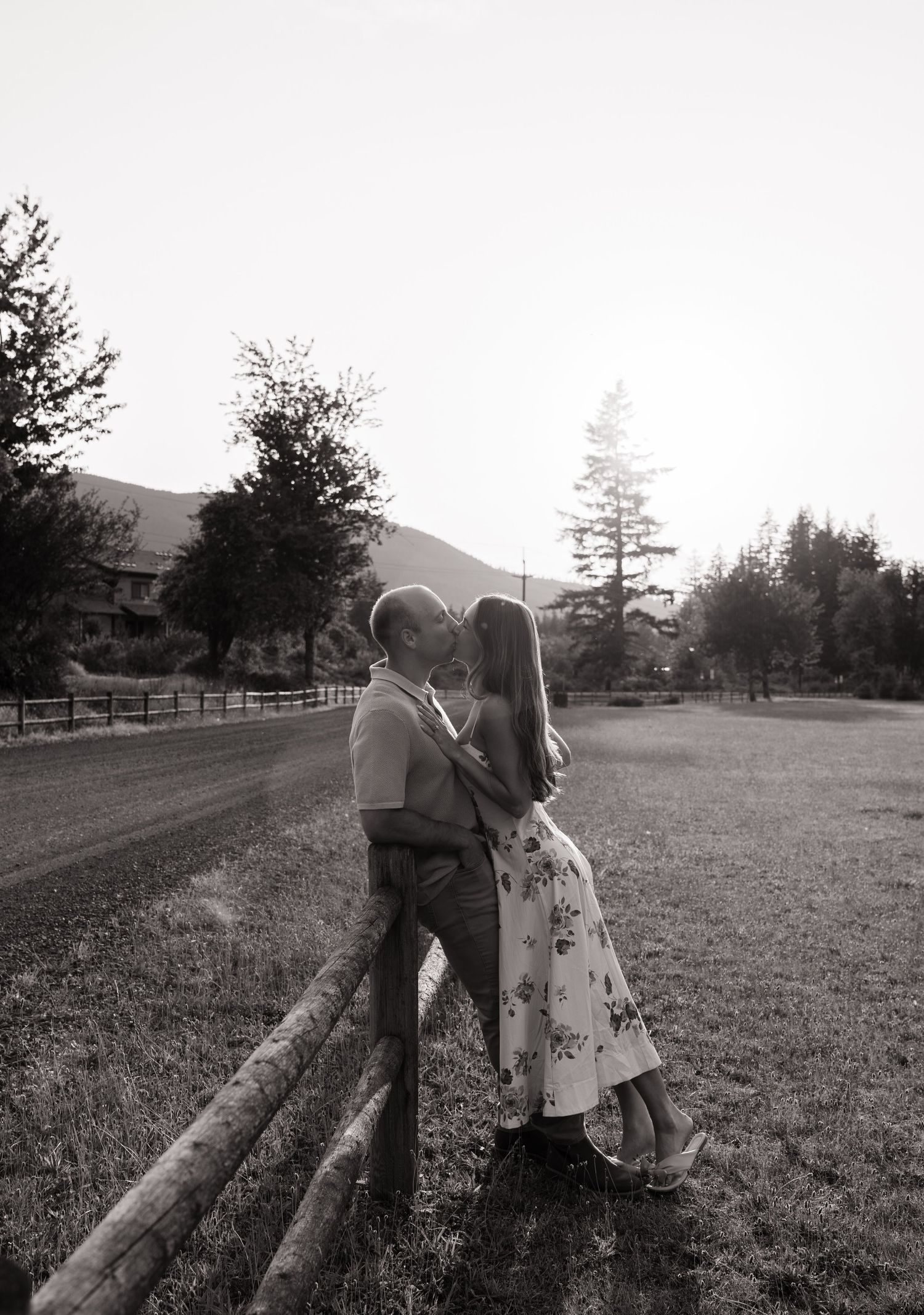 Black and white photo of a couple kissing against a rustic wooden fence with trees and mountains behind them.