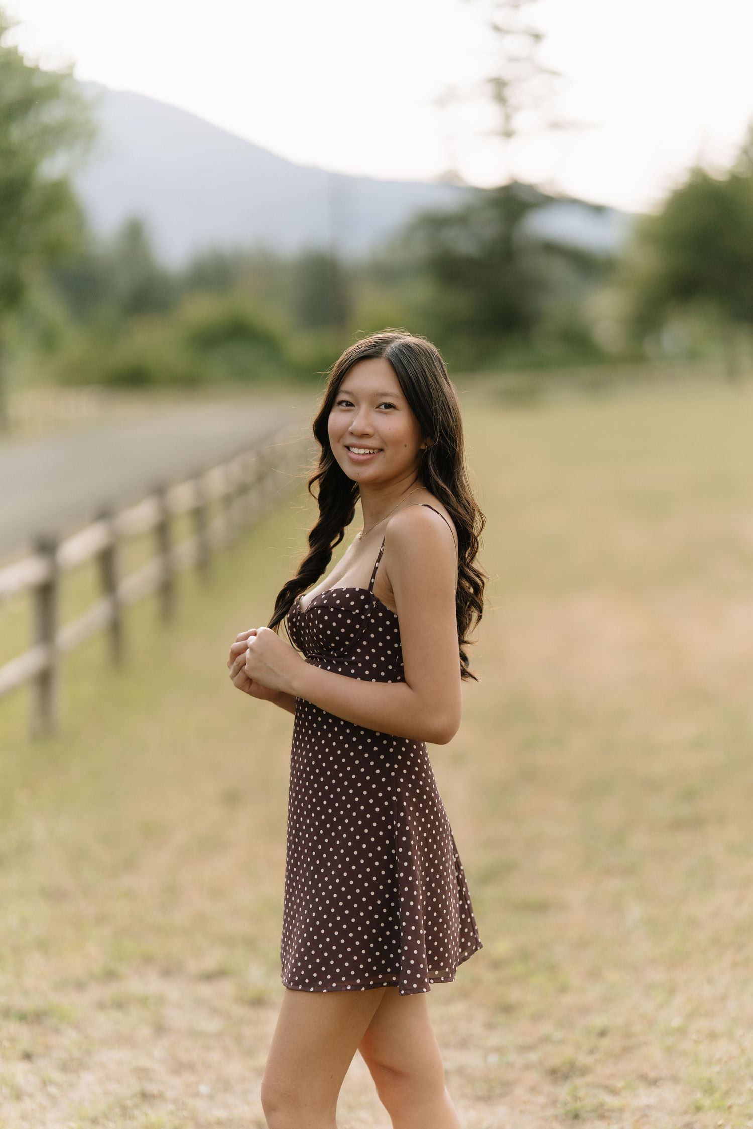 Smiling young woman in a brown polka dot dress stands in an open field with a wooden fence and mountain in the background.
