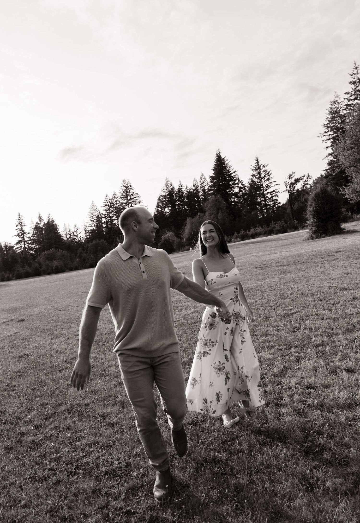 A black and white photo of a smiling couple holding hands and walking through a grassy field with evergreen trees behind them.