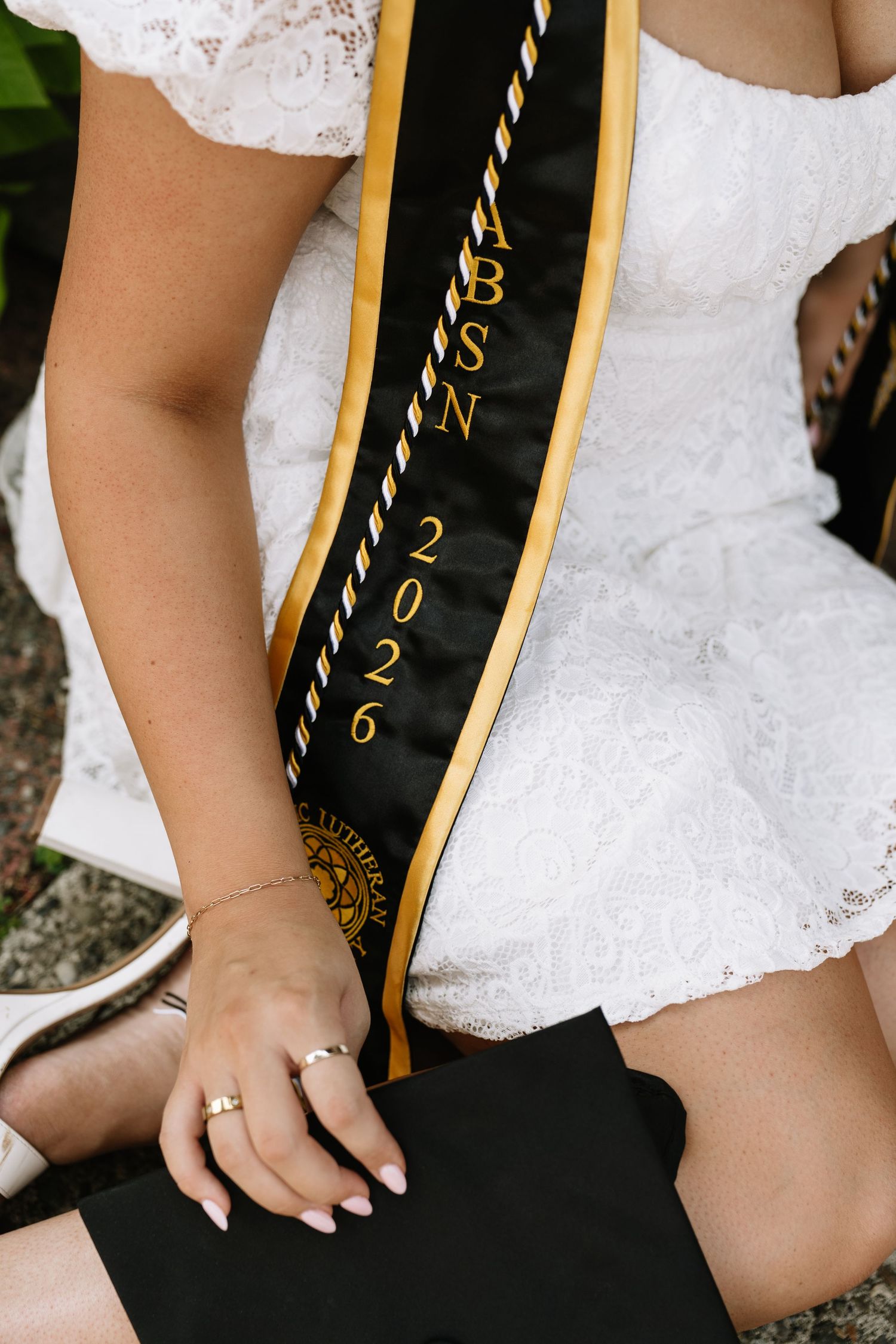 A woman in a white lace dress kneels with a black cap, wearing a black and gold 2026 graduation sash and rings on her hand.