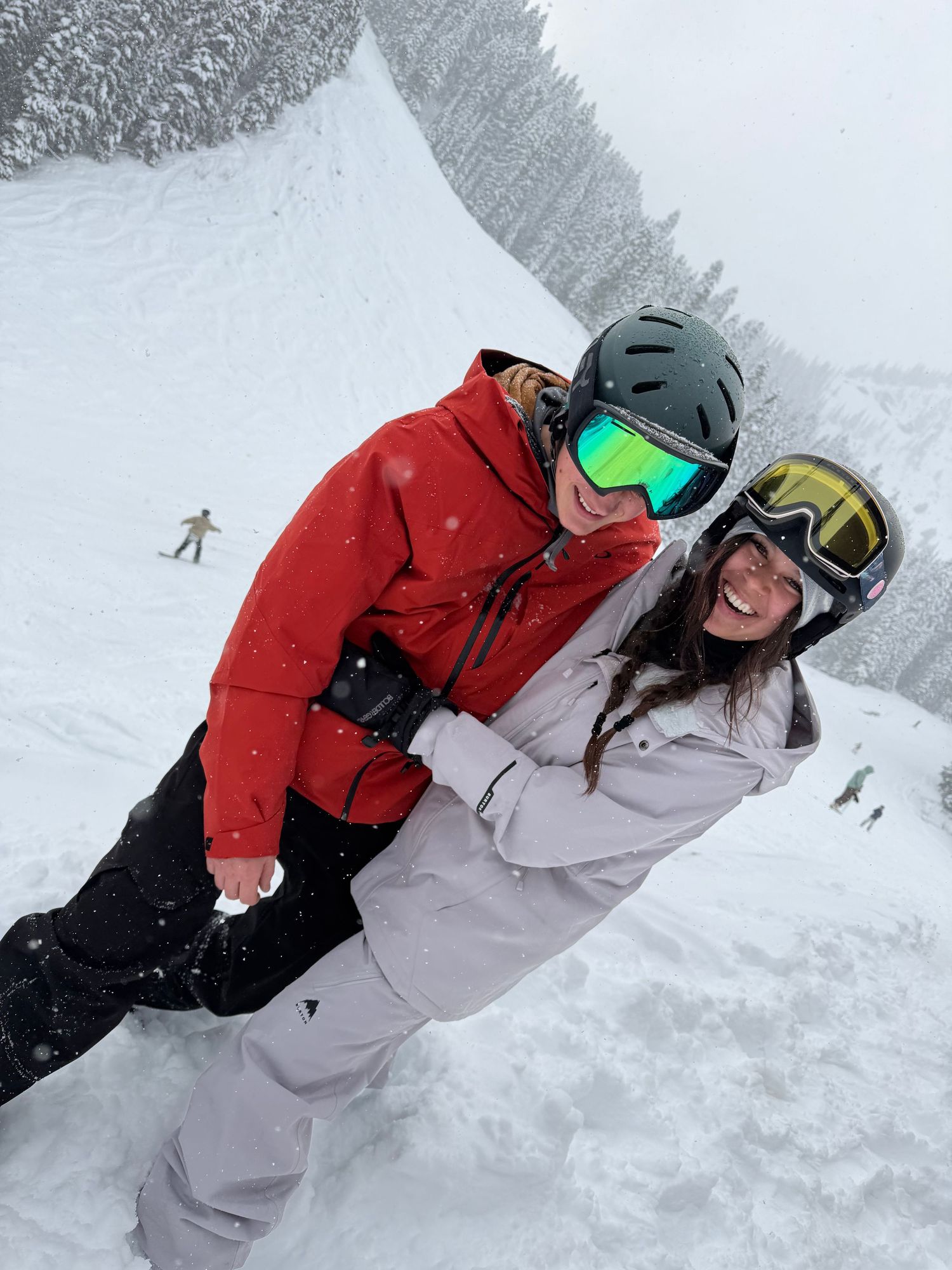 Two smiling snowboarders in goggles and helmets hug on a snowy mountain slope surrounded by snow-covered evergreen trees.