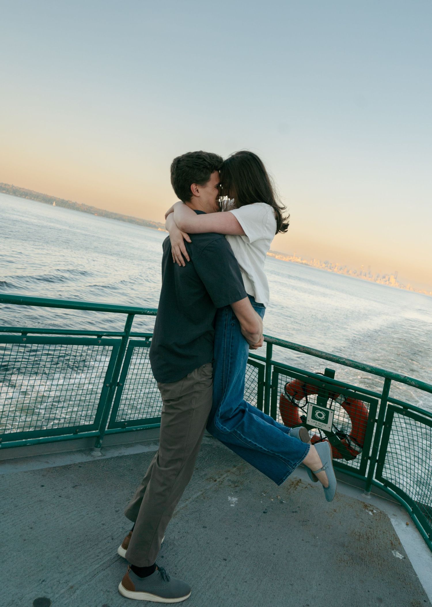 Man lifts and hugs a woman in a white tee and jeans on a ferry deck as the sun sets over calm water in a romantic engagement moment.
