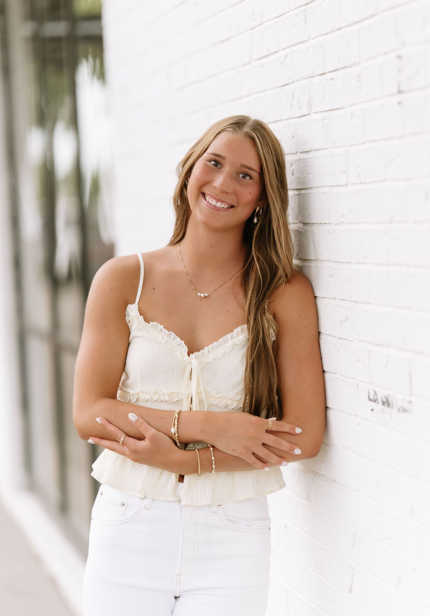 Smiling young woman in a cream ruffled top and white jeans leaning against a white brick wall.