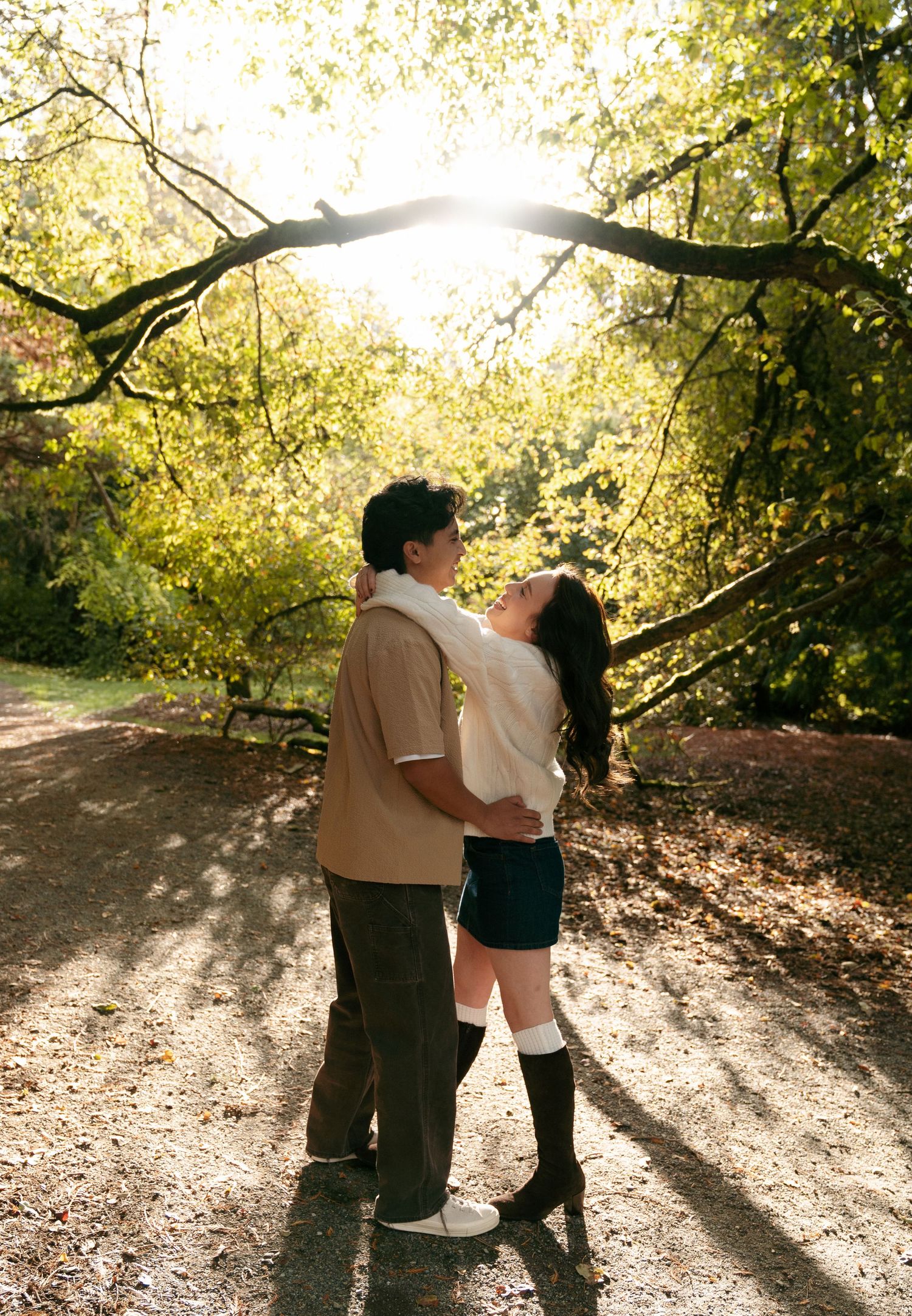 A couple embraces on a tree-lined path as golden sunlight streams through the leaves of an arching branch overhead.