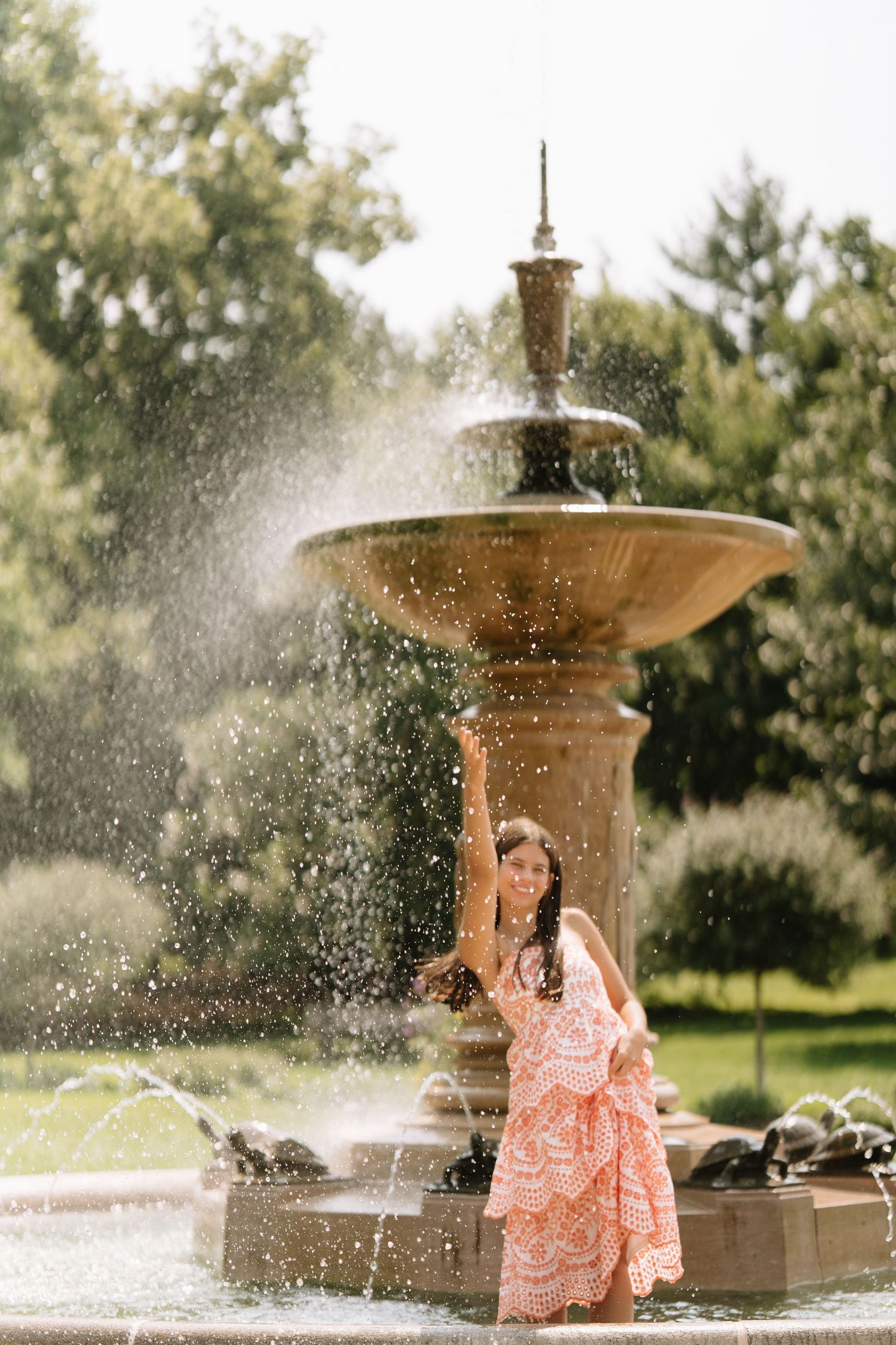 Woman in a coral lace dress raising one arm beside a stone fountain with water spraying in a sunlit garden.