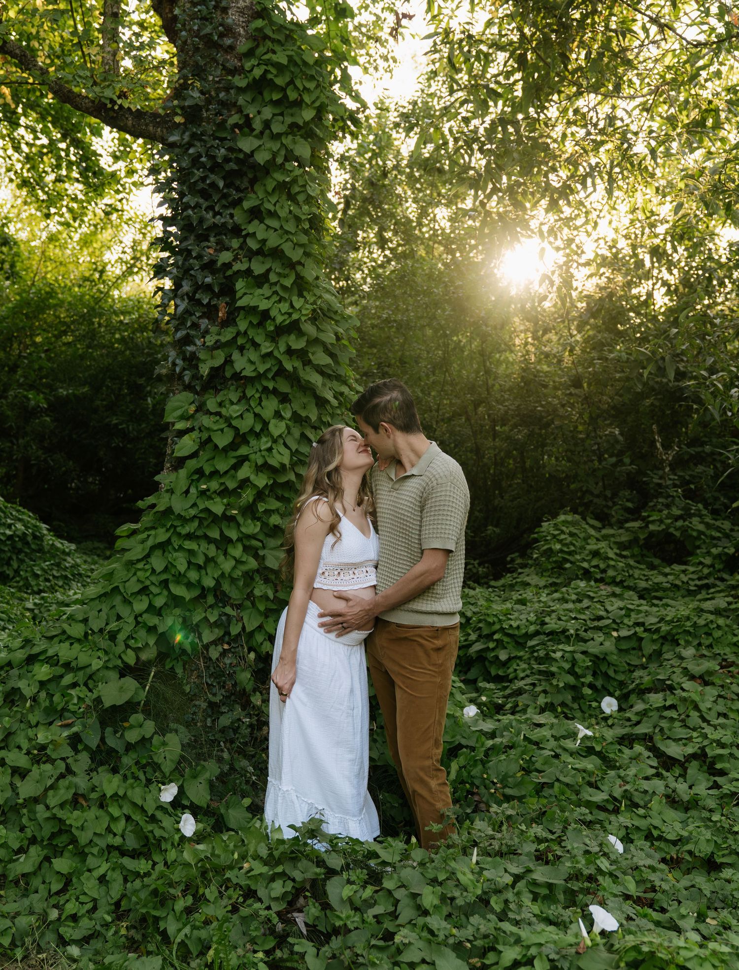 An engaged couple embraces for a kiss beneath an ivy-covered tree in a lush green garden with golden sunlight filtering through.