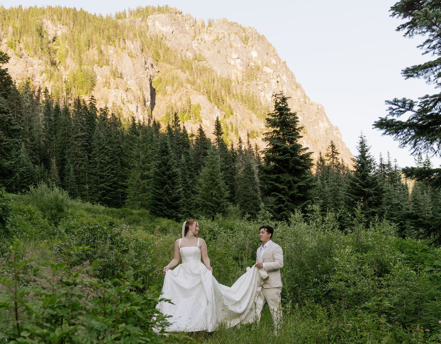 Bride and groom in a wedding gown and tan suit hold the flowing skirt in a grassy valley beneath a rocky mountain.