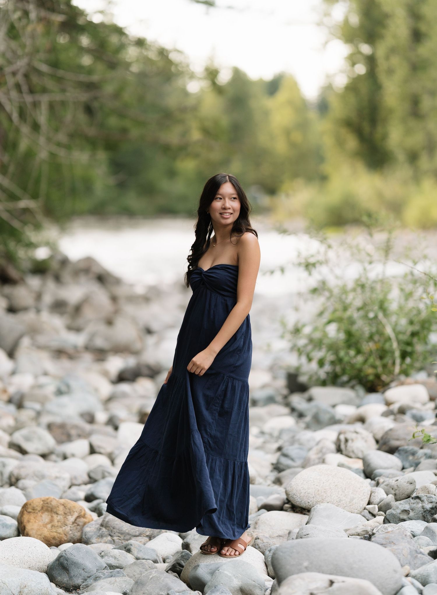 Woman in a flowing navy maxi dress standing on smooth river rocks with trees and water in the background.