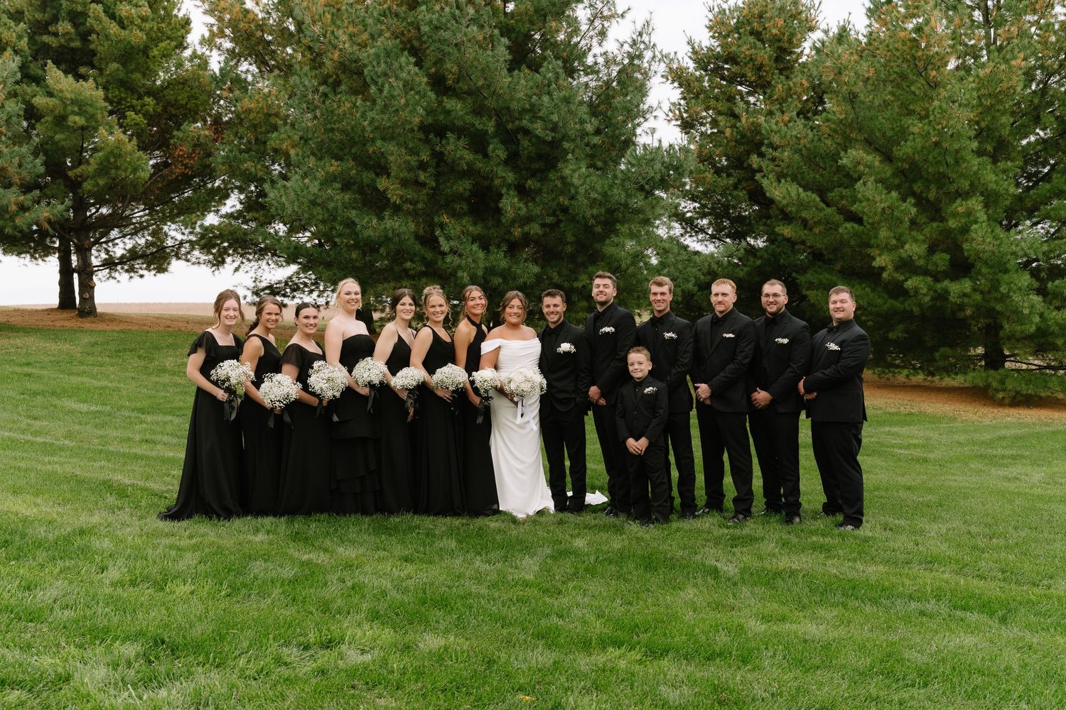 Bridesmaids in black dresses and groomsmen in dark suits pose with a bride in white on a green lawn near tall evergreen trees.