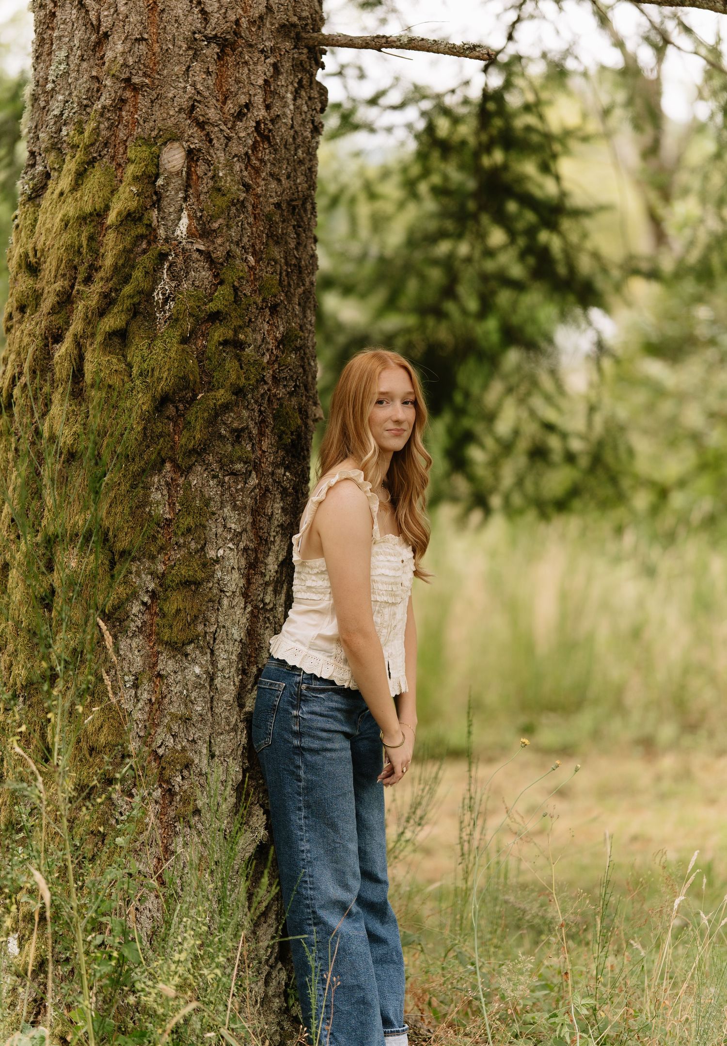 Young woman with long strawberry-blonde hair in a cream top and jeans leans against a mossy tree trunk in a grassy field.