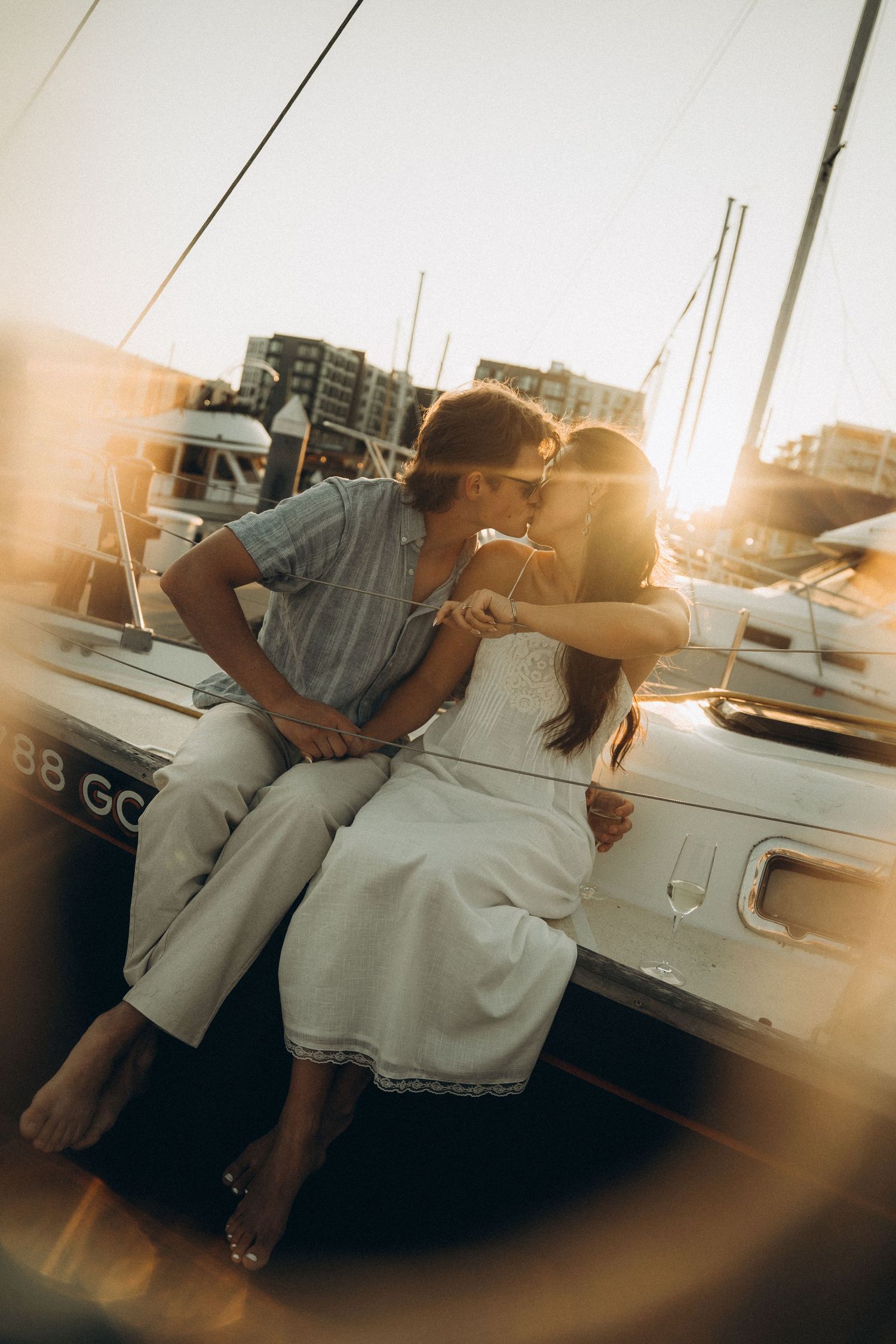 A couple kisses on the edge of a boat at a marina during a warm, glowing golden hour sunset.