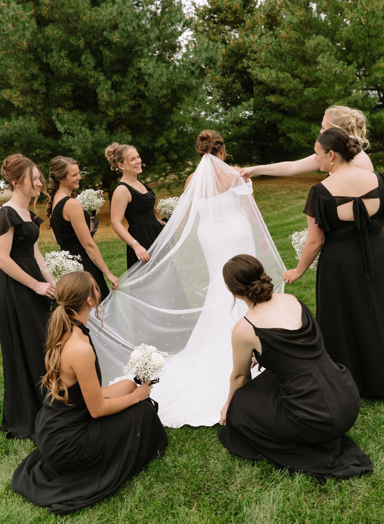 Bridesmaids in black dresses gather around a bride on the grass, adjusting her long flowing veil and holding baby's breath bouquets.
