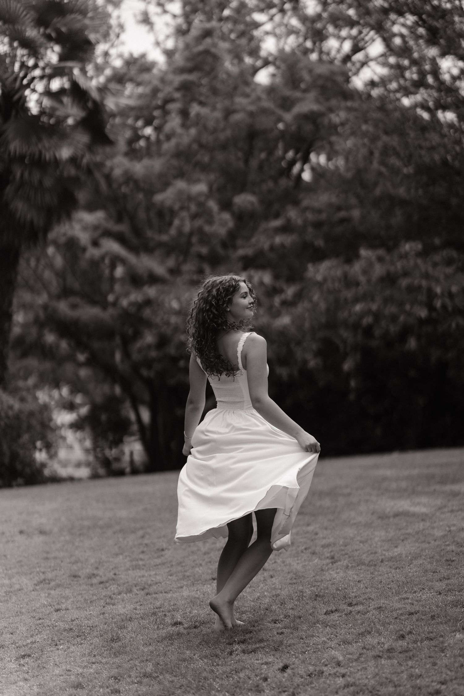 Black and white photo of a barefoot woman with curly hair twirling her white dress on a grassy lawn surrounded by trees.