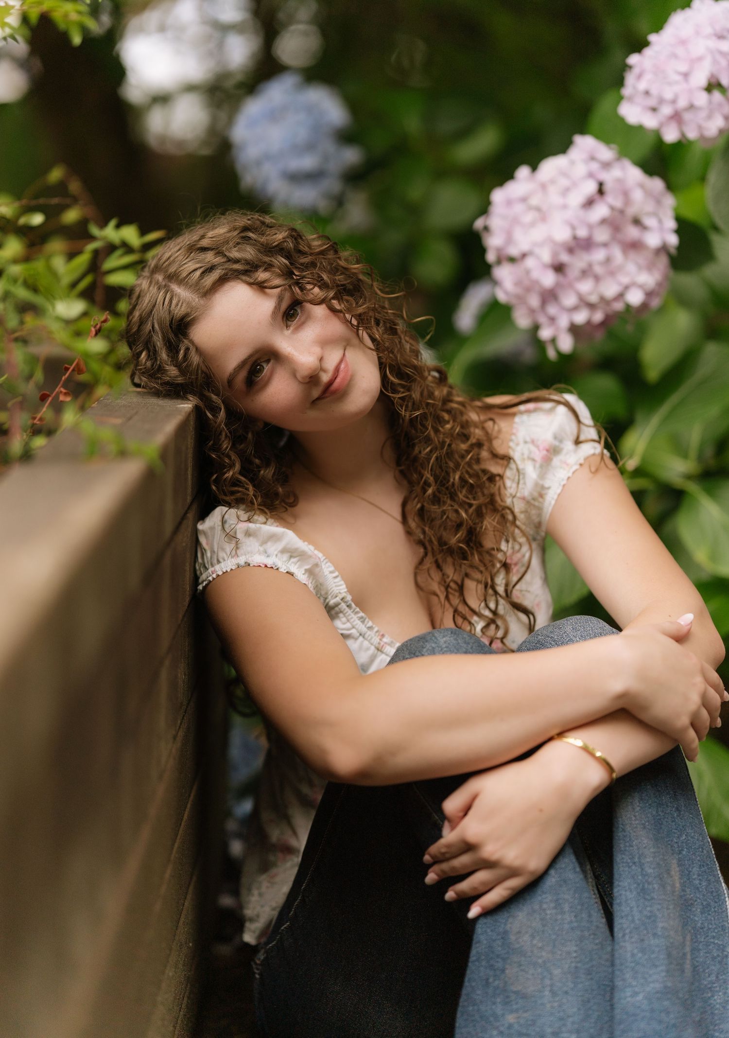 Young woman with long curly hair leans against a wooden wall, smiling beside pink hydrangeas in a lush garden portrait.