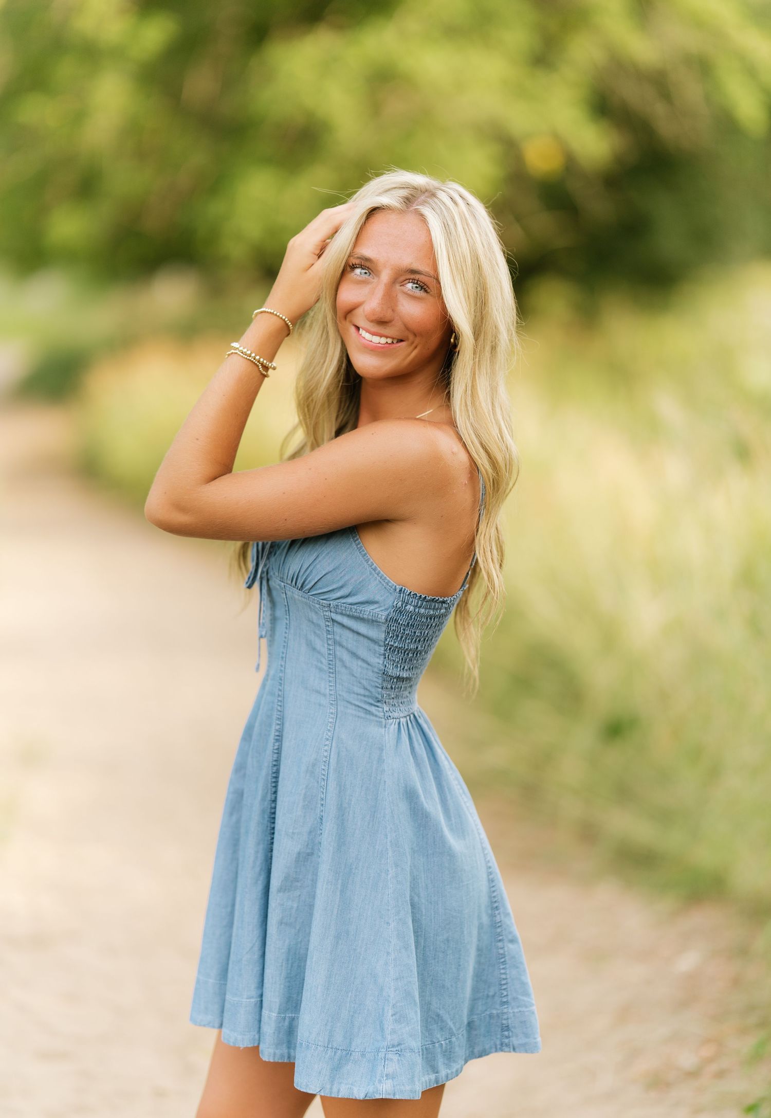 Blonde woman in a blue denim sundress smiling with hands in her hair on a sunlit country path.