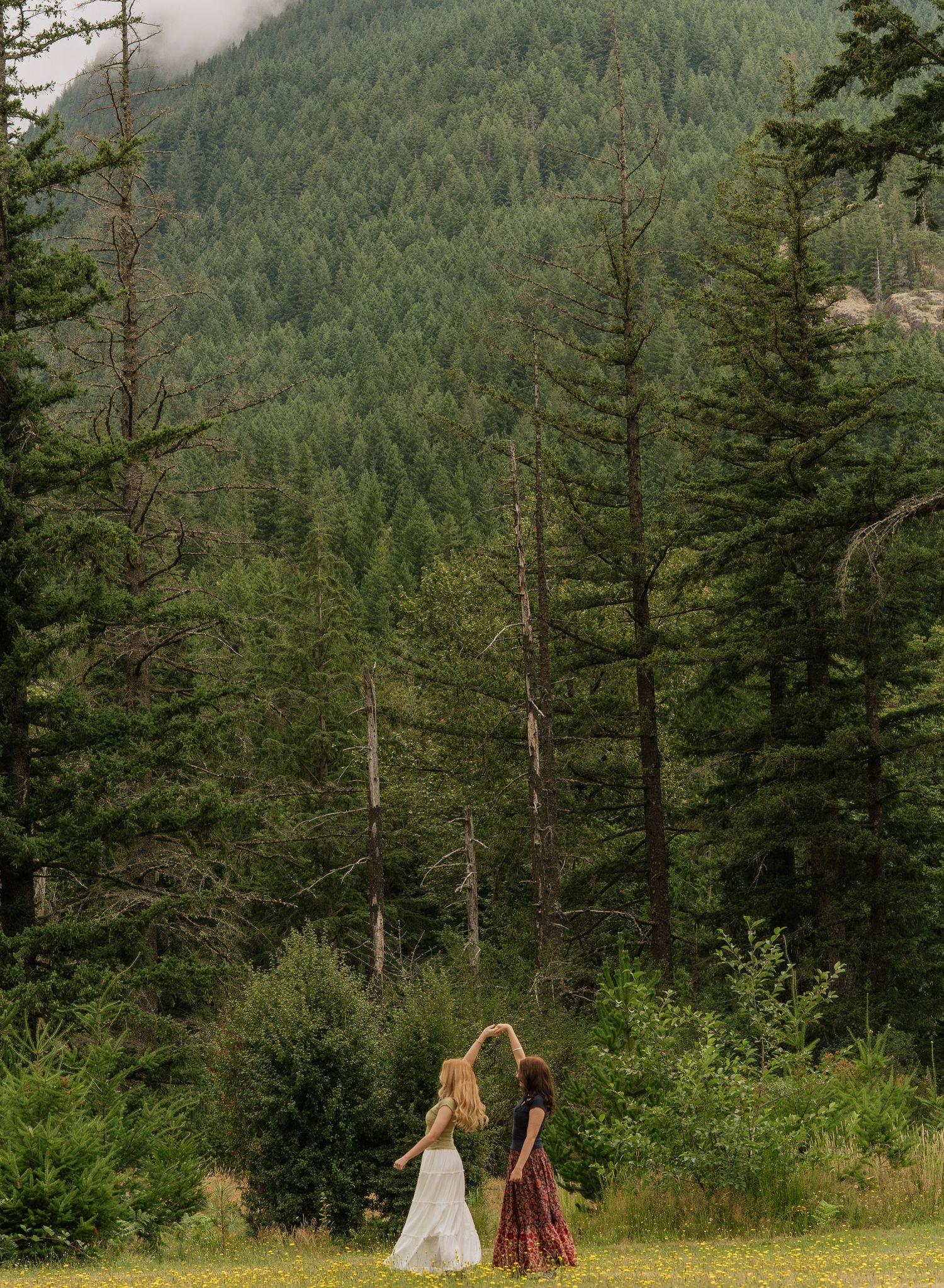 Two women in flowing dresses dancing in a grassy meadow with a forested mountain behind them.