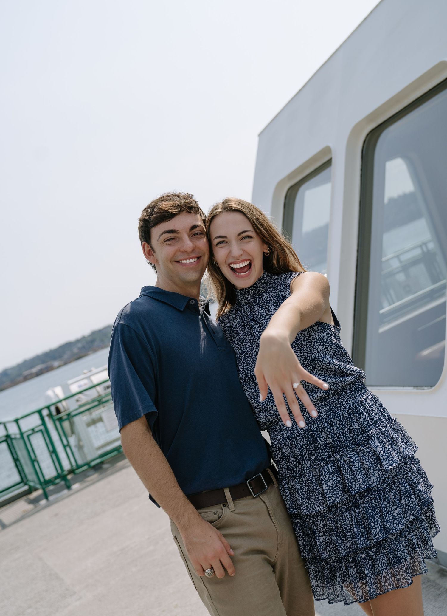 A smiling couple poses together on a ferry dock while the woman playfully extends her hand toward the camera.