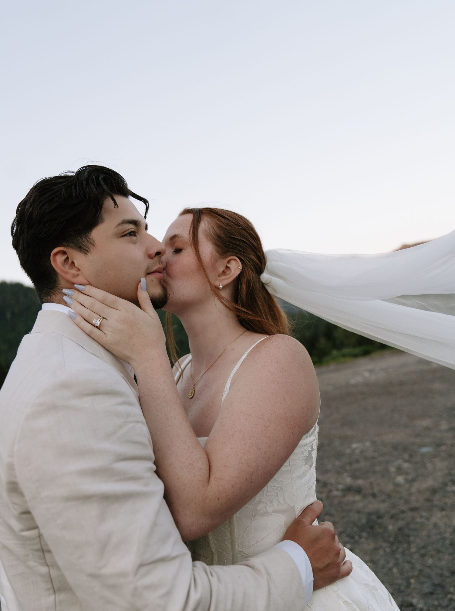 Close-up of a bride and groom kissing as her long veil blows in the wind, with a ring visible on her hand.