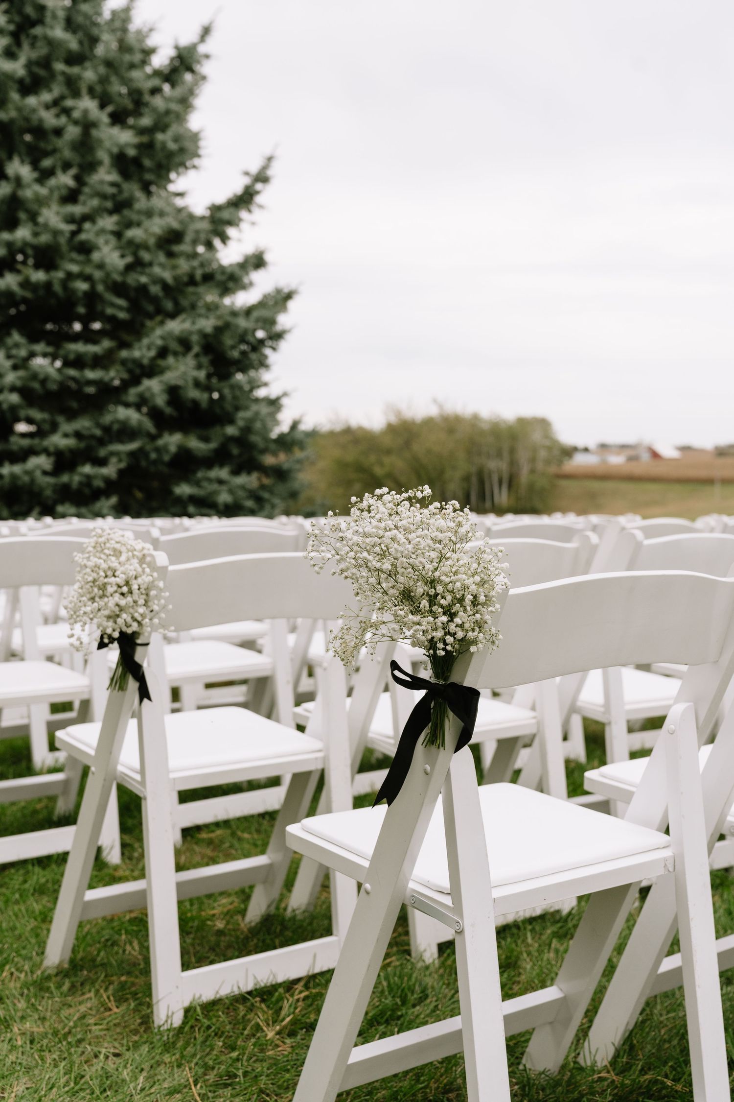 White folding chairs with baby's breath bouquets and black ribbons line a grassy outdoor wedding ceremony space near evergreen trees.