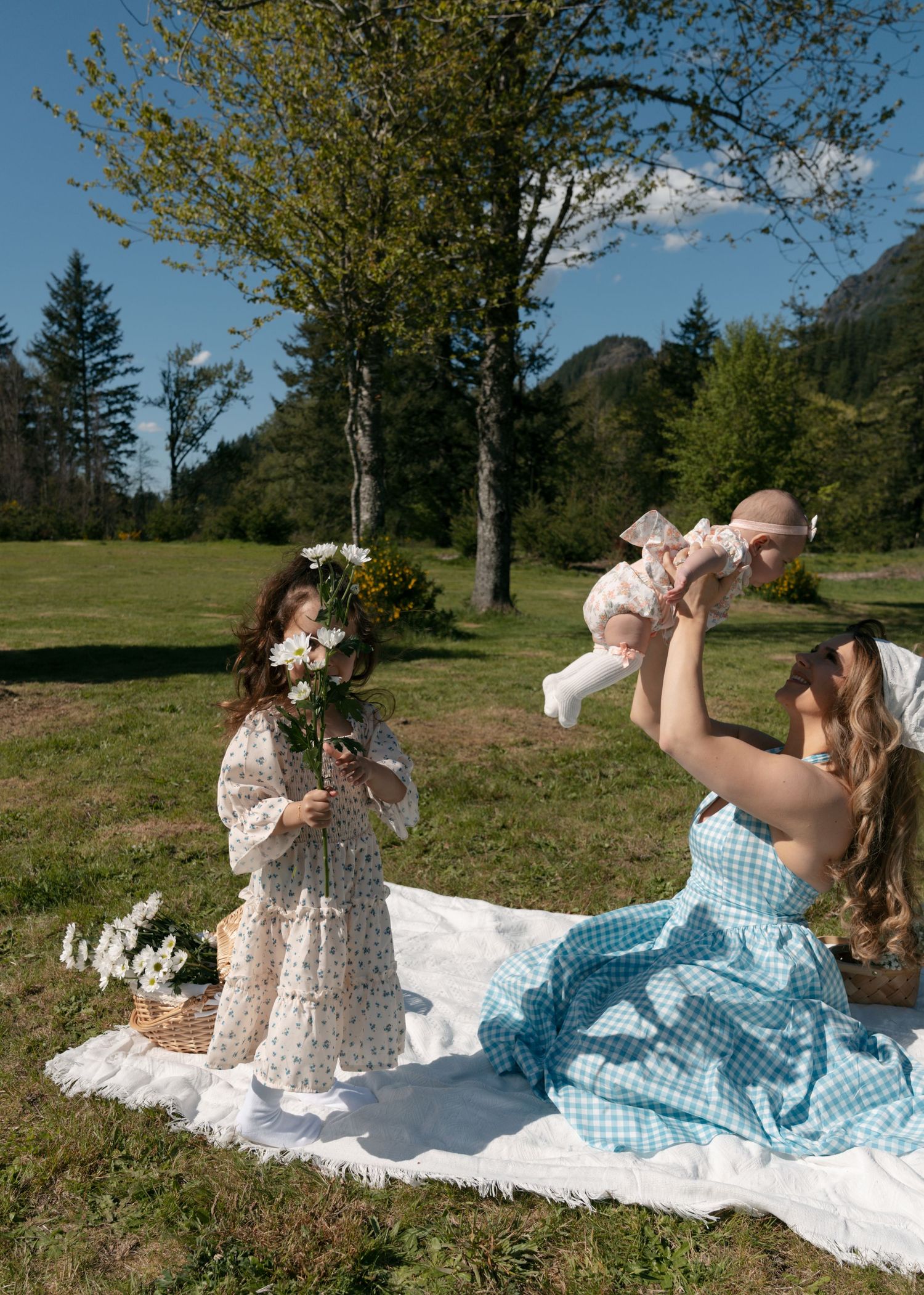 A mother in a blue gingham dress lifts her baby overhead on a picnic blanket as a young girl holds white flowers in a mountain meadow.