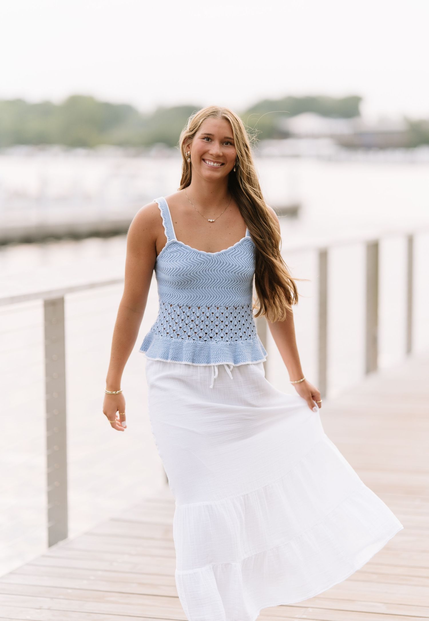 Smiling young woman with long hair in a blue crochet top and white skirt stands on a waterfront boardwalk by a railing.