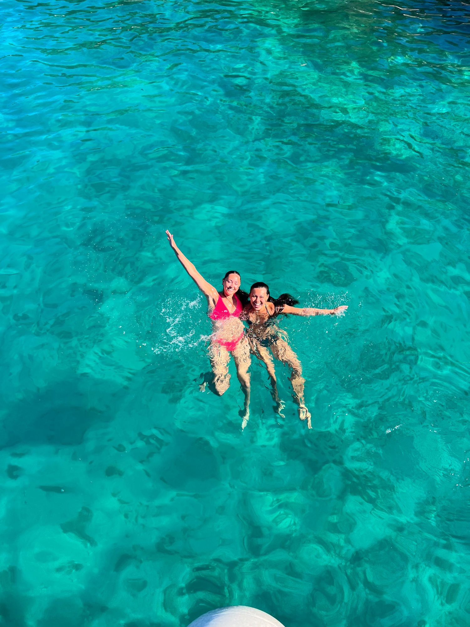Two smiling women in bikinis swim and wave their arms in clear turquoise water, photographed from above.