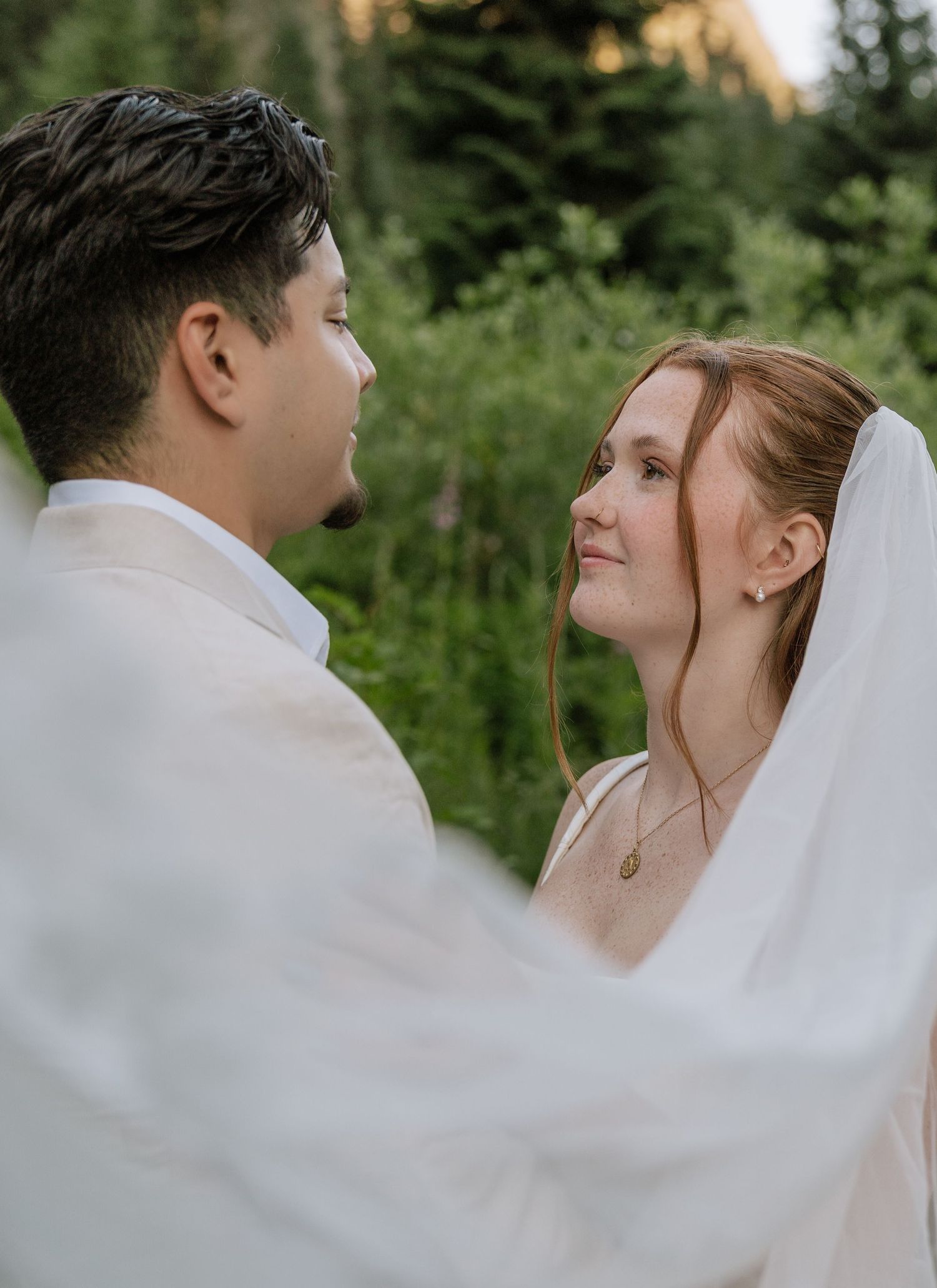 Bride with red hair and a sheer veil gazes lovingly at her groom in a lush green forest setting.