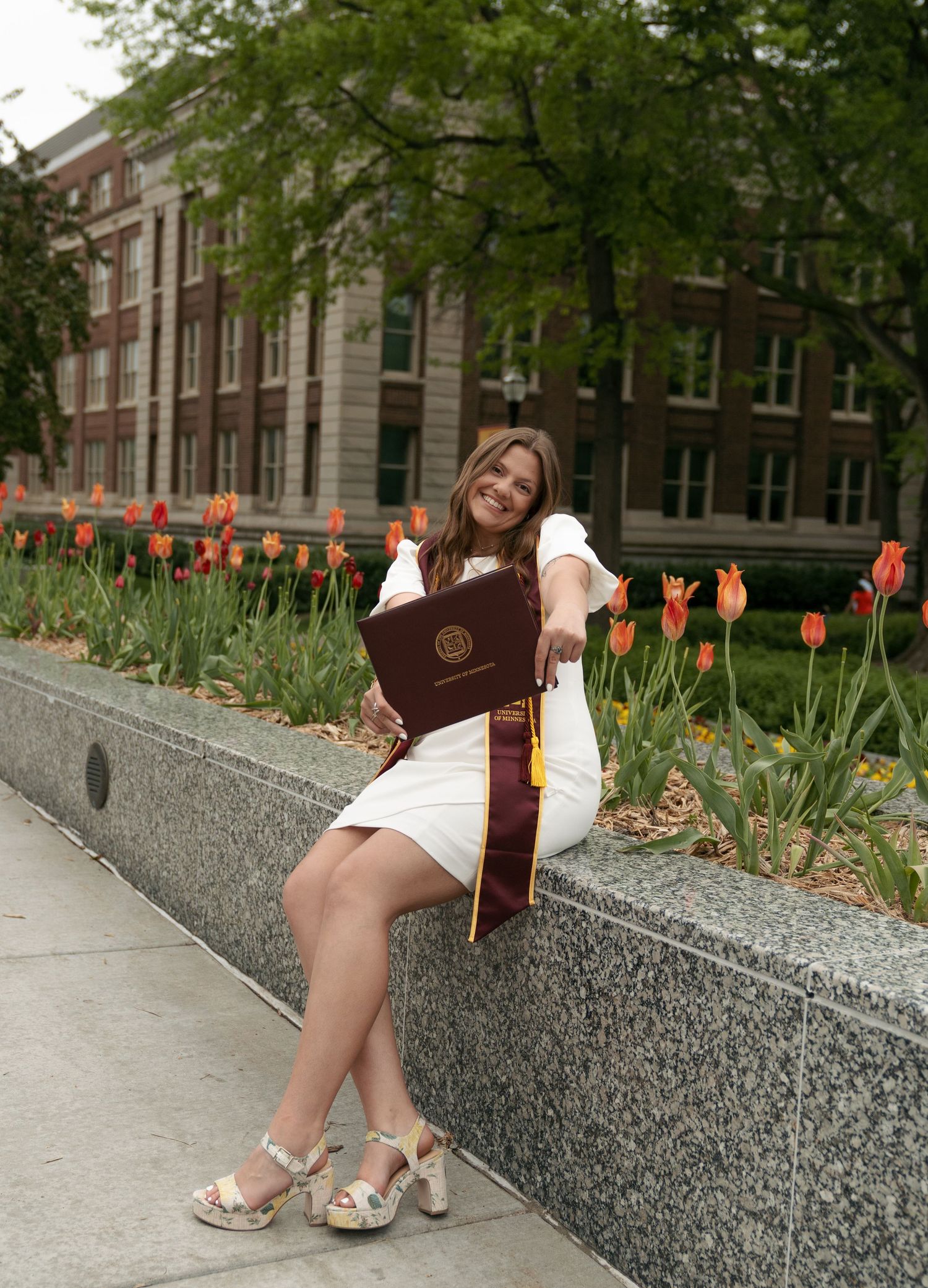 Smiling graduate in a white dress and platform sandals sits on a stone ledge holding a maroon diploma cover beside blooming orange tulips.