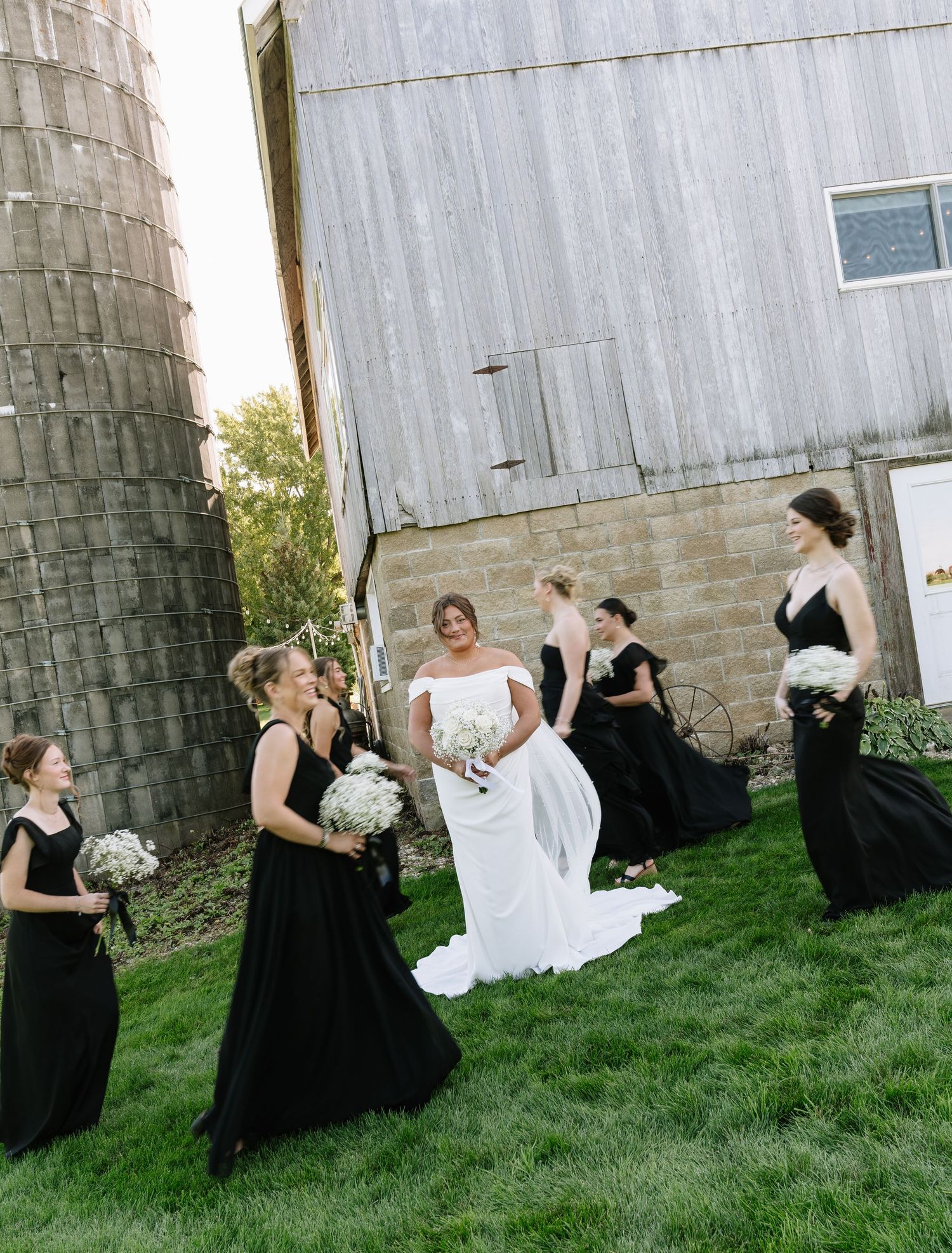 A bride in a white gown stands on a lawn with bridesmaids in black dresses beside a rustic barn and stone silo.