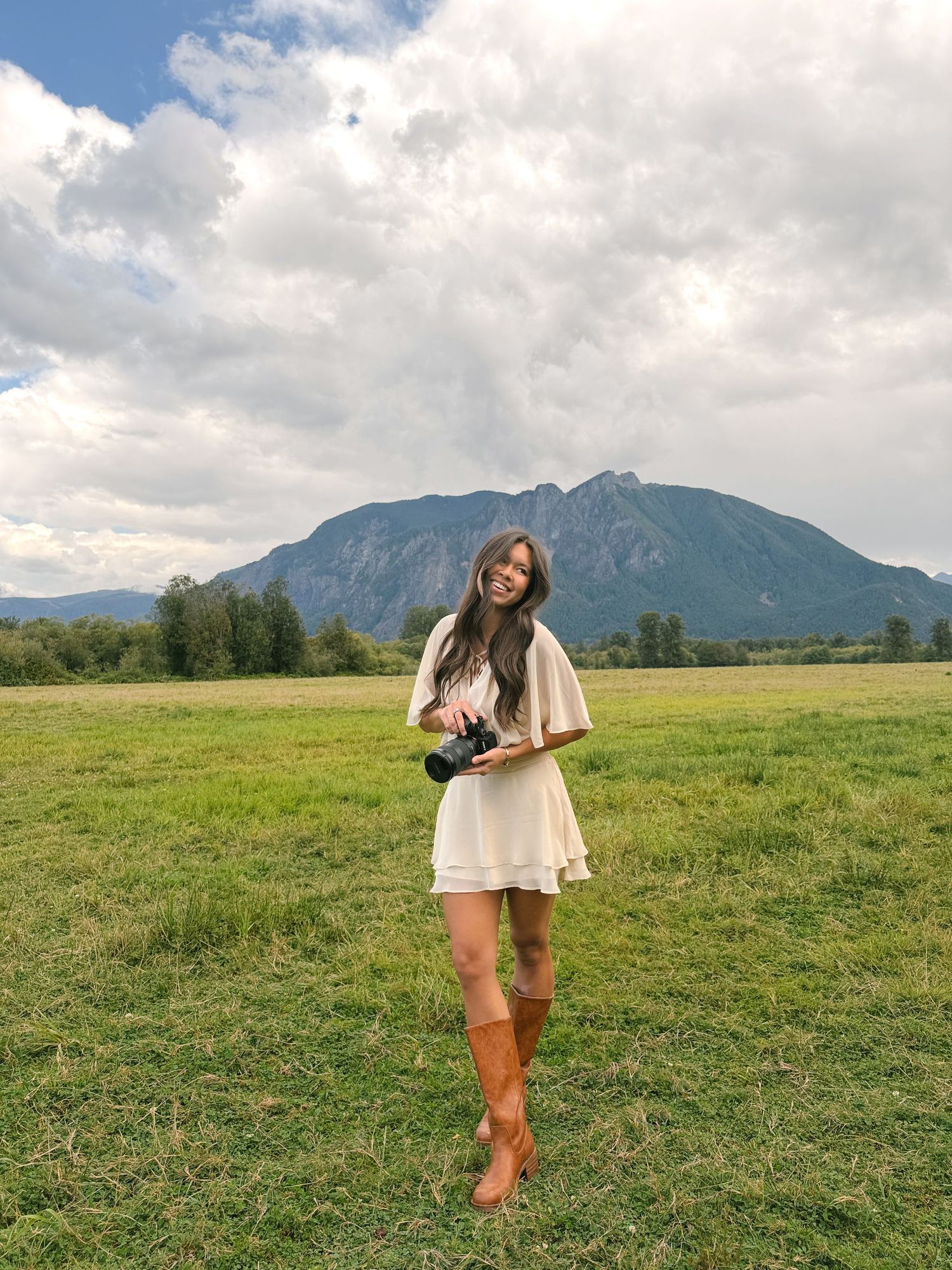A smiling woman in a cream dress and brown boots holds a camera in a grassy field with a mountain behind her.