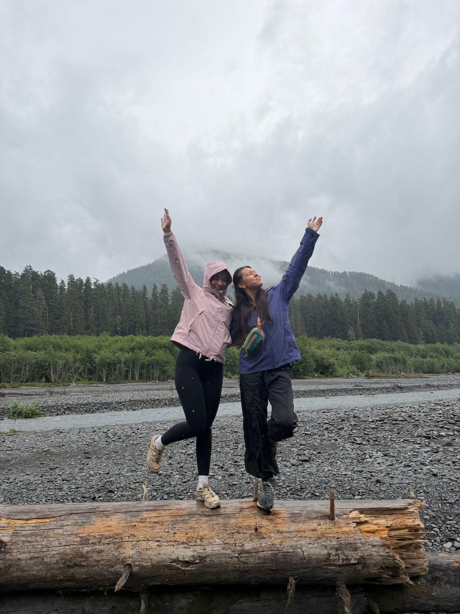 Two women in rain jackets pose with raised arms on a driftwood log along a gravel riverbank with forested mountains behind.