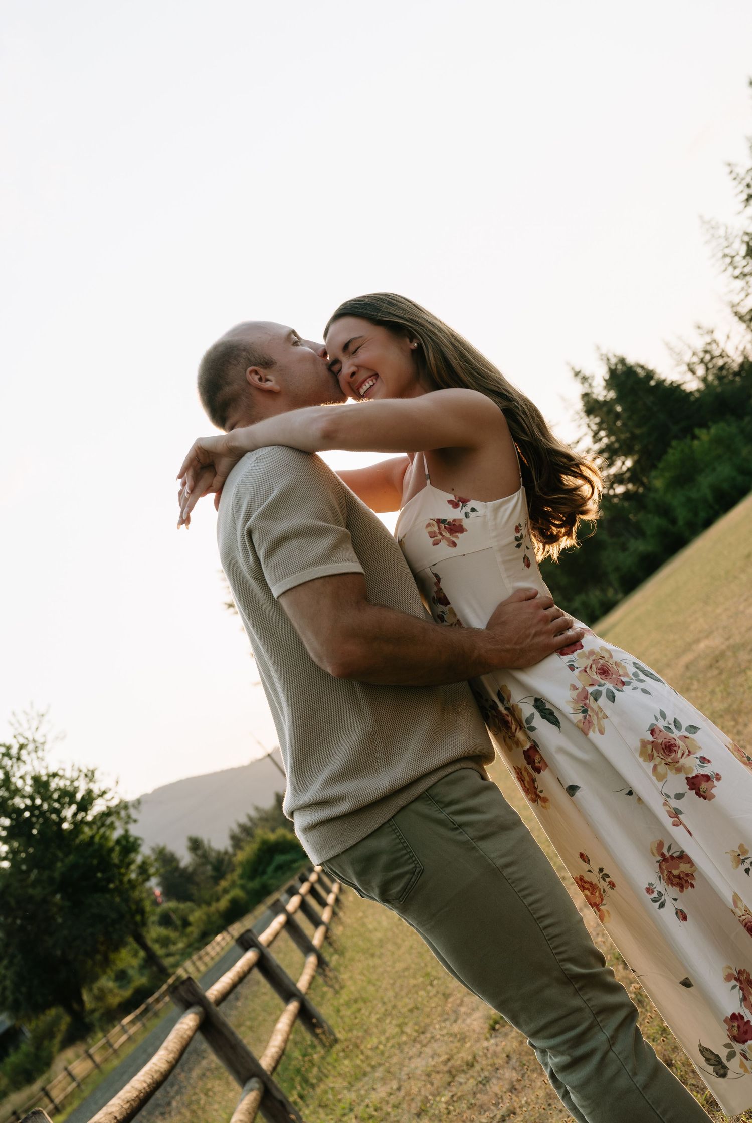 A couple embraces and kisses in a sunlit grassy field beside a wooden fence, the woman wearing a floral dress with mountains behind them.