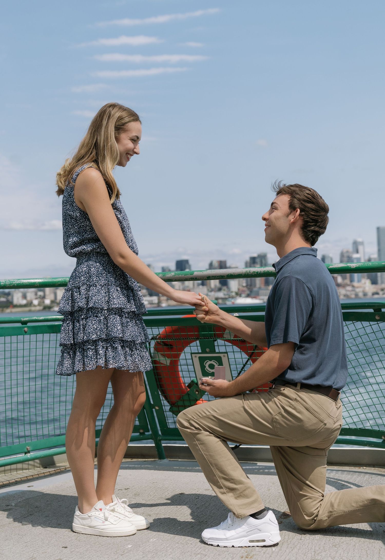 Man kneeling on one knee proposes to a smiling woman in a floral dress on a ferry deck with a city skyline behind.