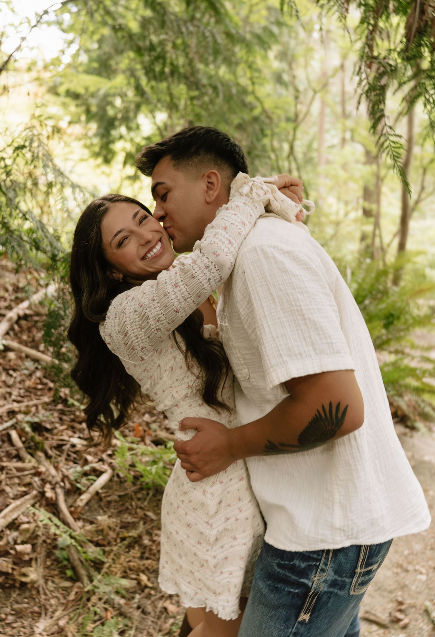 Smiling woman in a white eyelet dress embraces a man in a white shirt as he kisses her cheek in a sunlit forest.