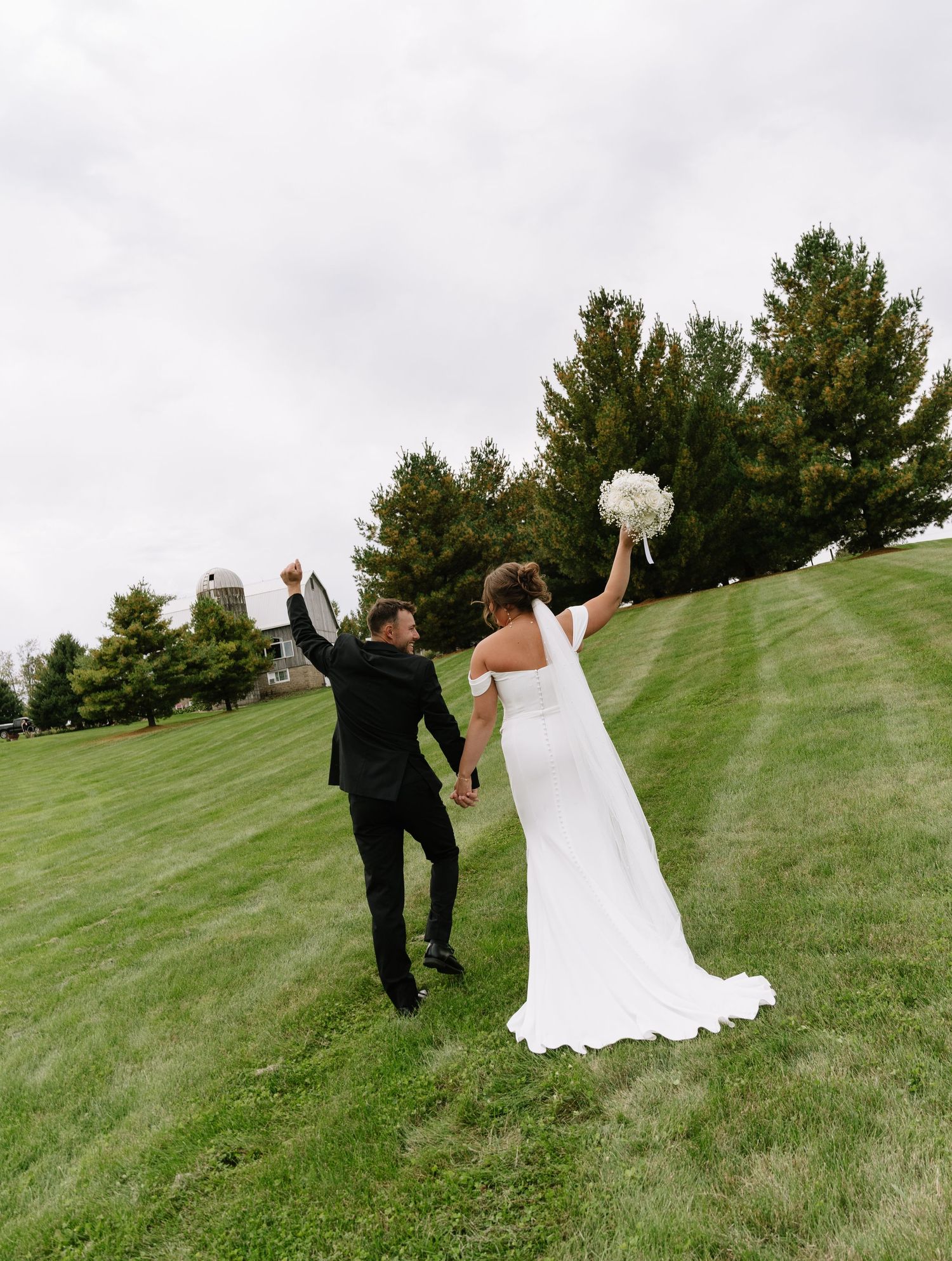 Bride in a white gown and groom in a black suit walk hand in hand up a grassy hill, celebrating with raised arms and a white bouquet.