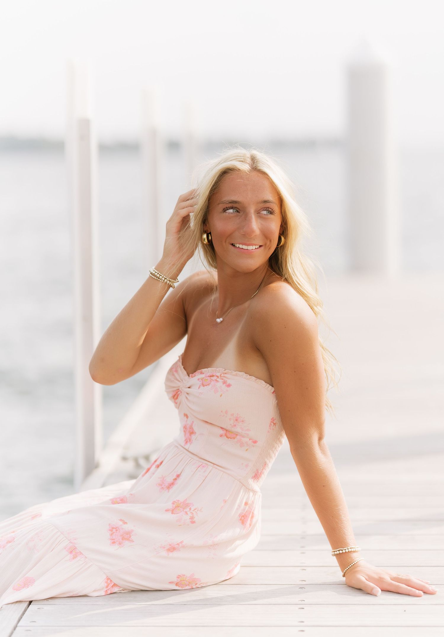 Smiling blonde woman in a pink floral sundress sits on a wooden dock by calm water, touching her hair in soft natural light.