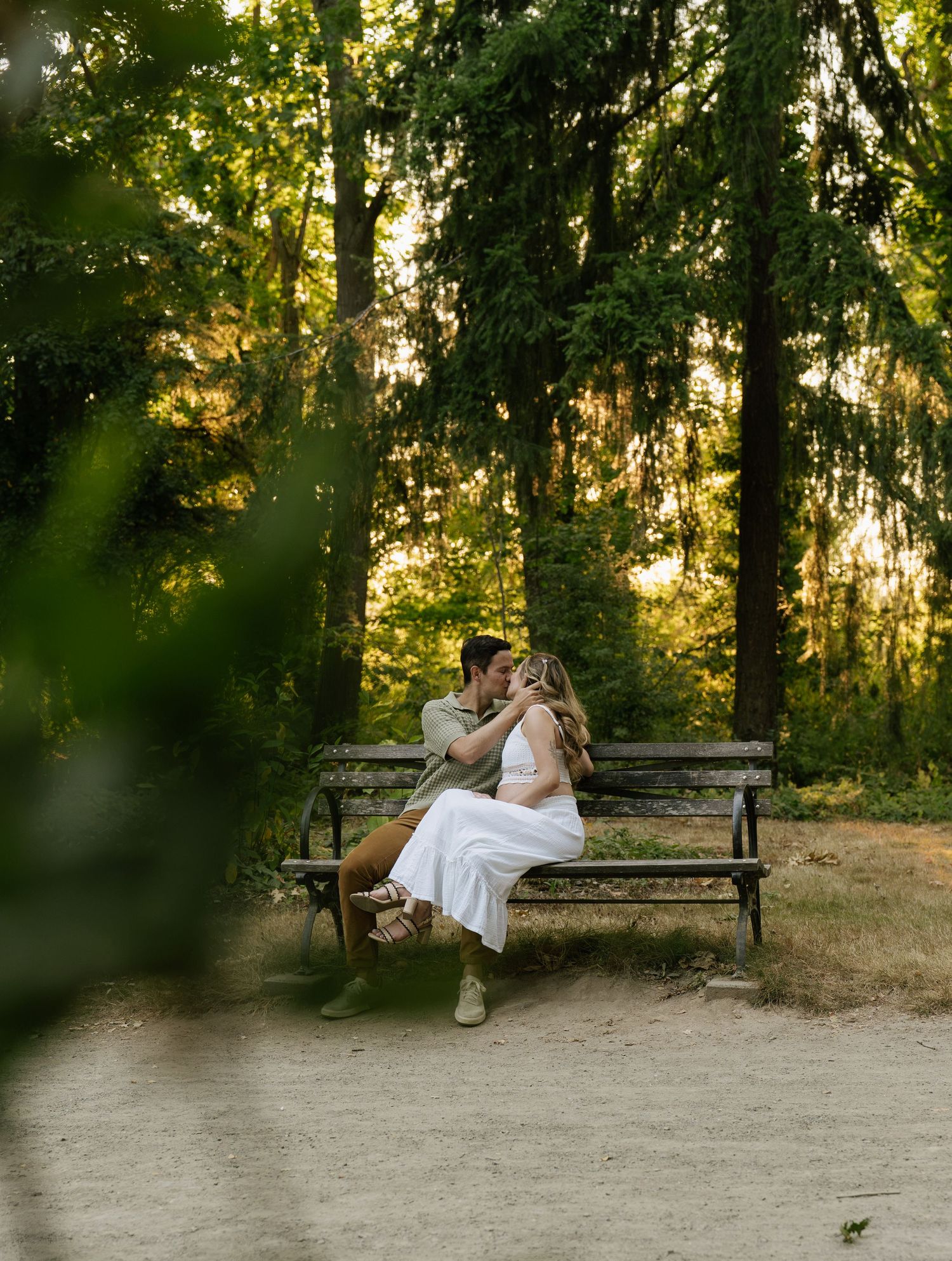 A couple kisses on a park bench surrounded by green trees in warm, golden forest light.
