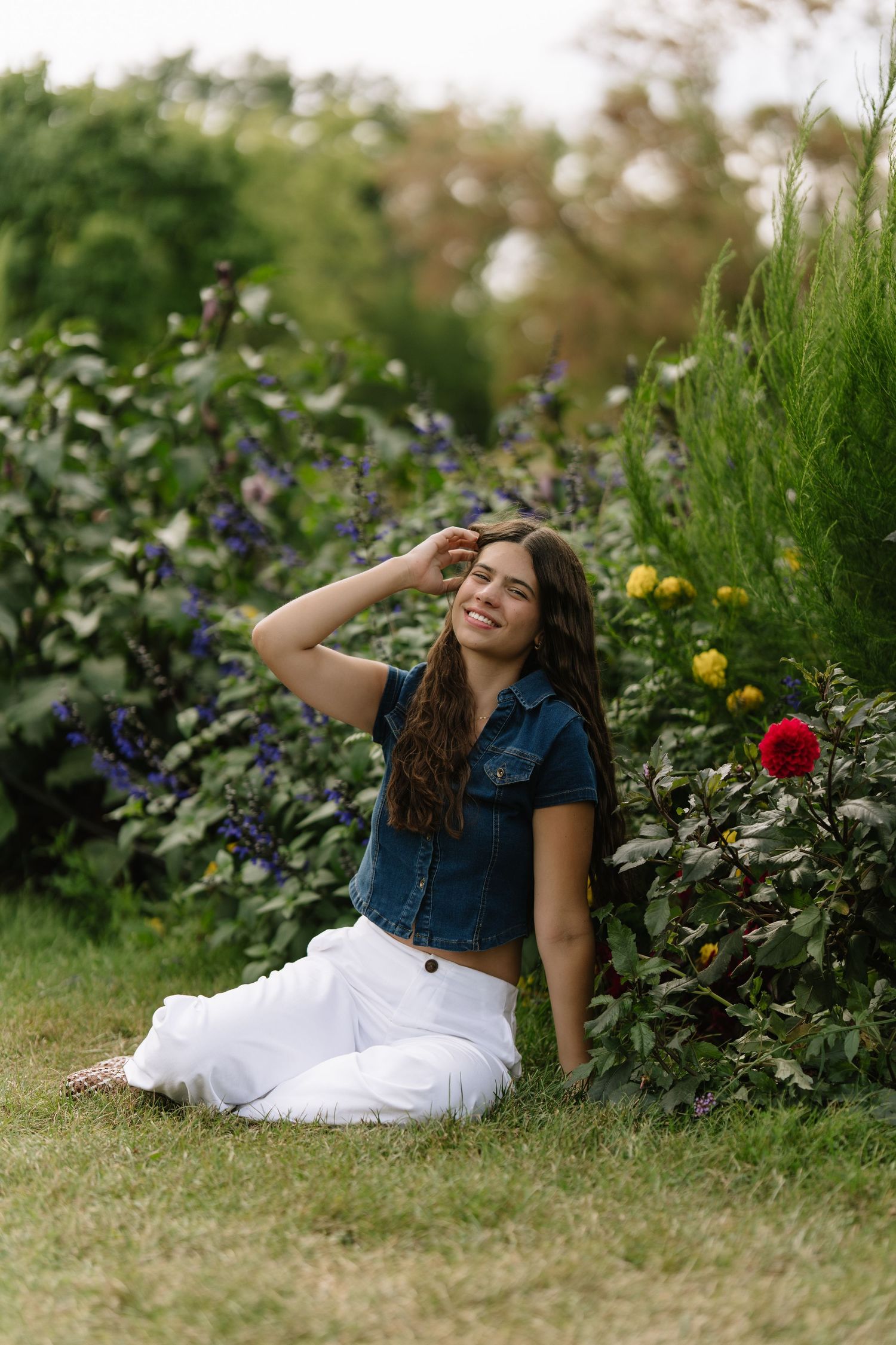 Smiling young woman in a denim top and white pants sits on grass beside purple flowers and red and yellow blooms in a garden.