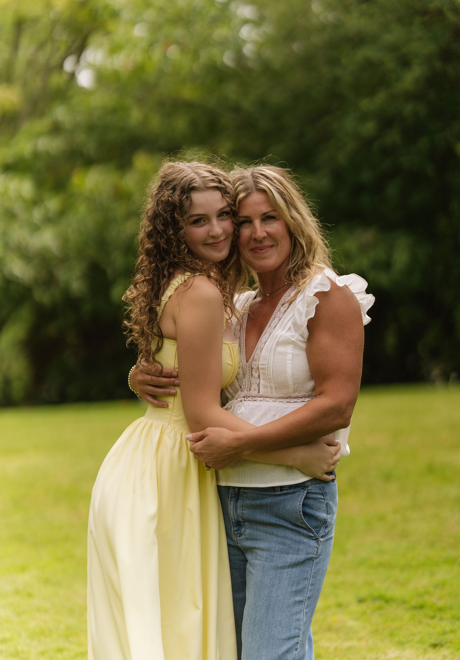 Smiling mother and daughter embracing outdoors on a green lawn, one in a yellow dress and one in jeans.