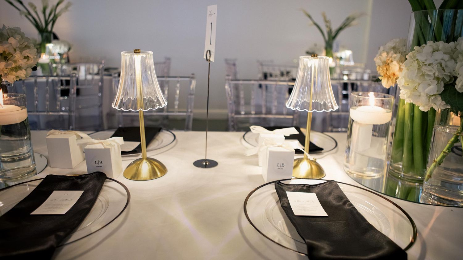Elegant wedding reception table with clear glass plates, black napkins, gold lamps, white candles, and floral centerpieces.