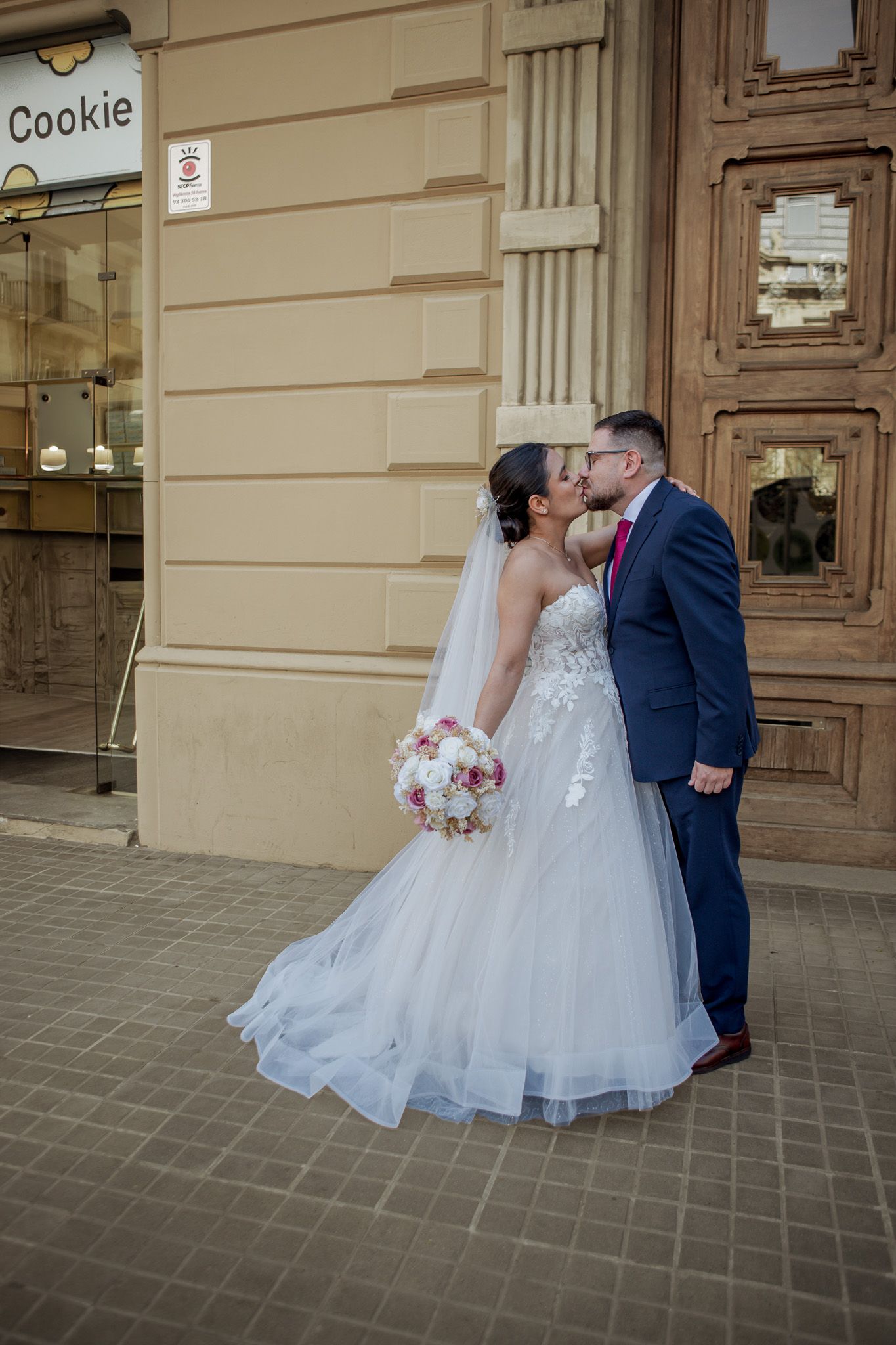 Bride in a lace tulle wedding gown holding a pink bouquet kisses her groom in a navy suit outside an elegant stone building.