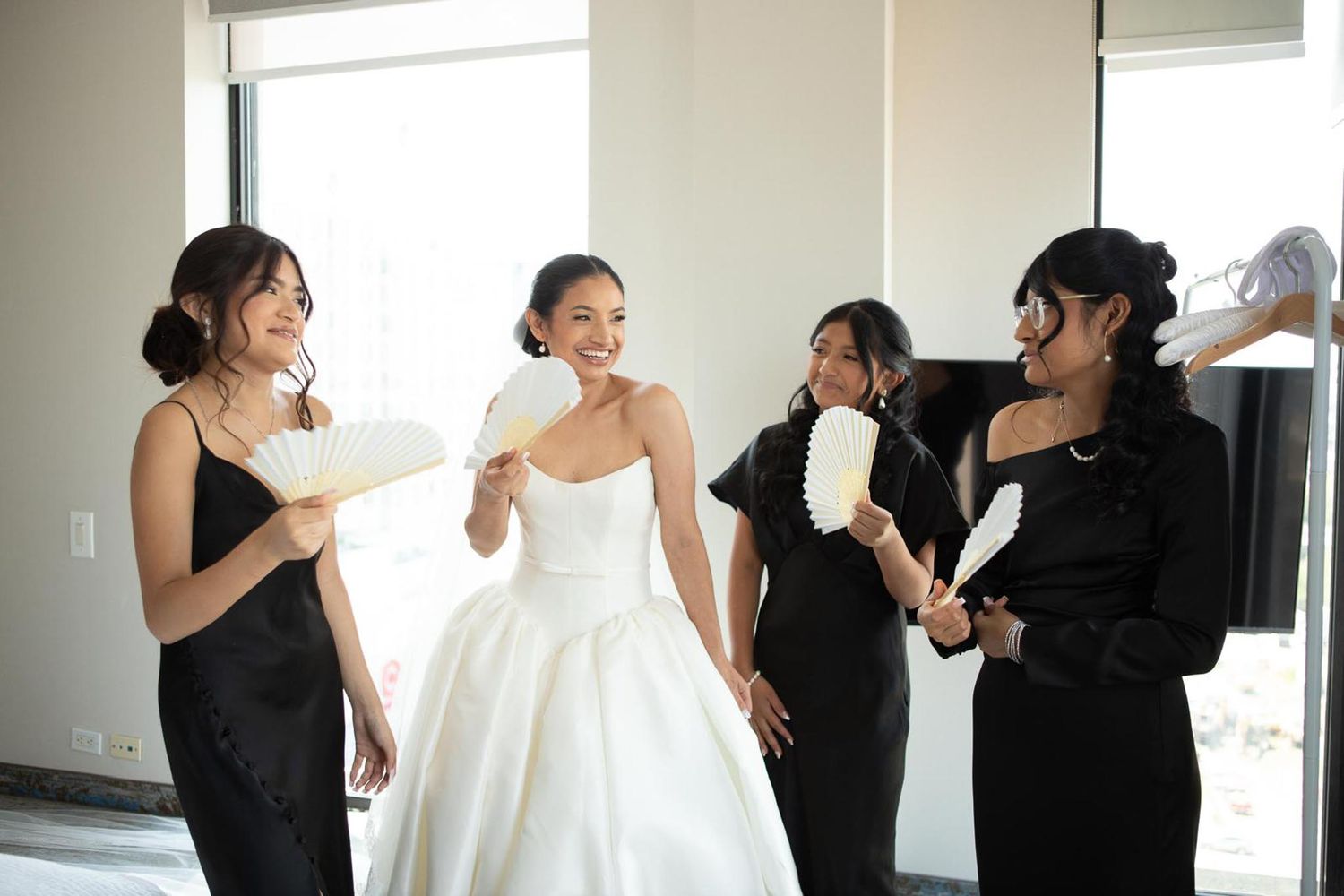 Bride in a strapless wedding gown laughs with bridesmaids in black dresses holding white hand fans in a bright room.