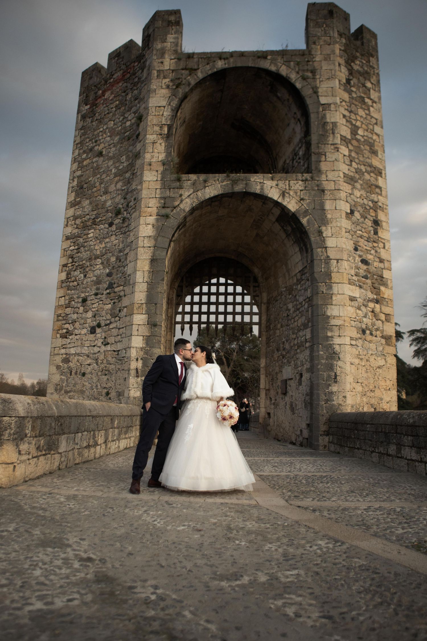 Novios abrazados frente al arco de una torre de piedra durante su sesión de boda. Besalú, España.